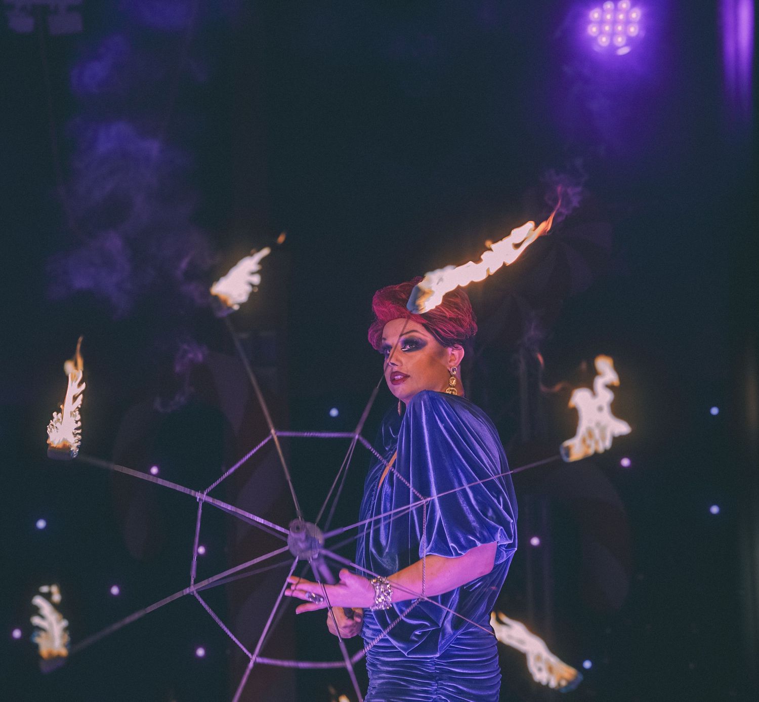 A performer in blue creates a dramatic display with fire props against a dark stage.
