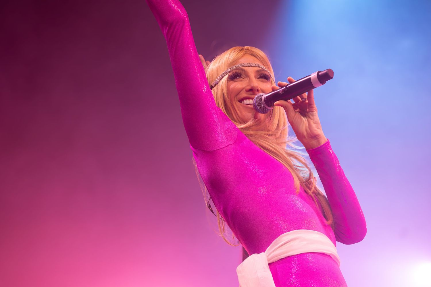 Concert performer in pink outfit singing energetically on stage with dramatic lighting effects.
