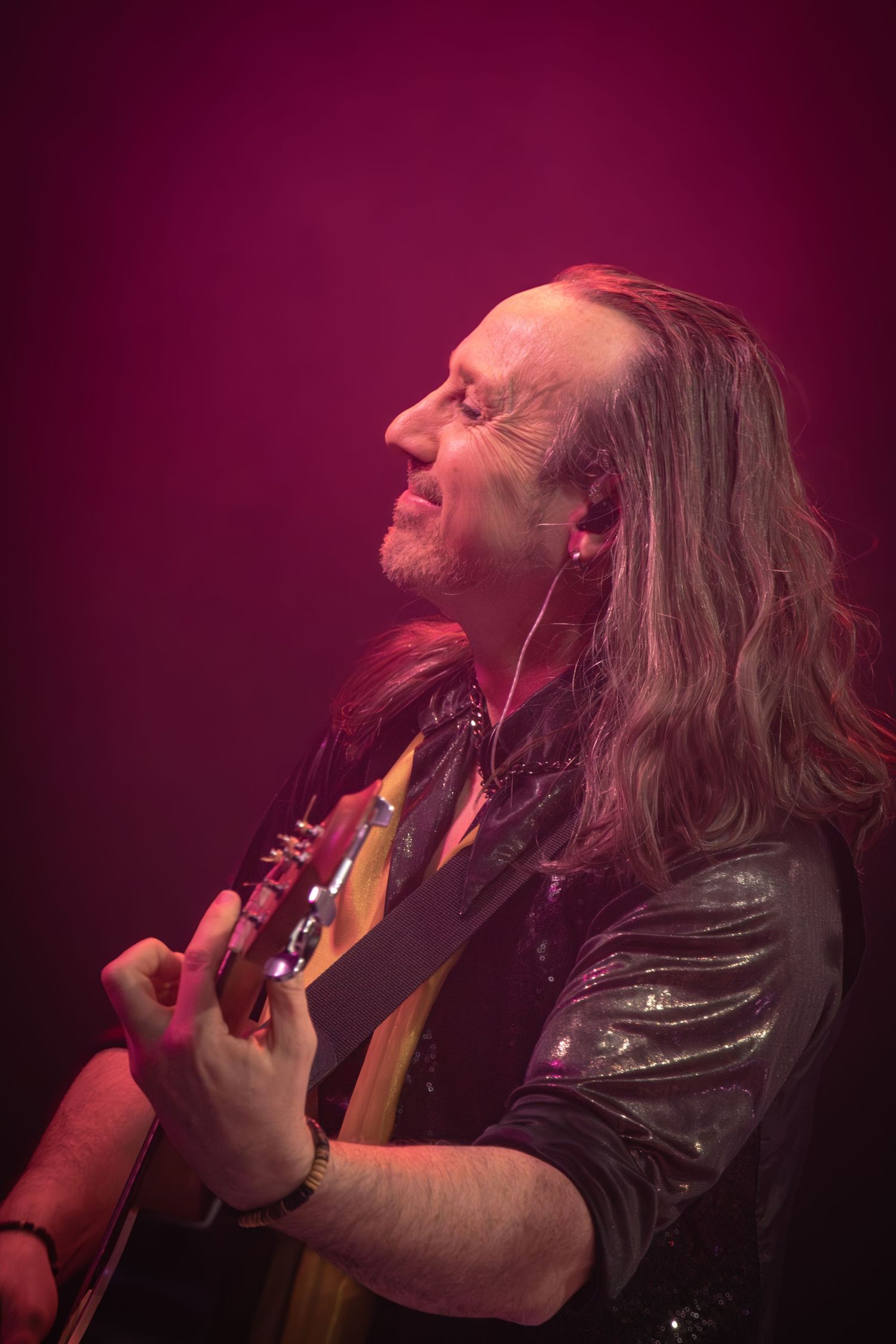 Musician with long hair playing guitar under moody red stage lighting.