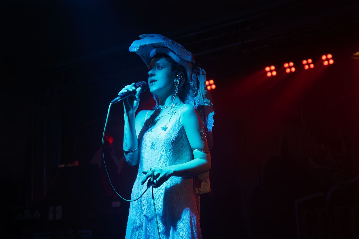A performer in a white dress sings passionately on stage under dramatic blue and red lighting effects.