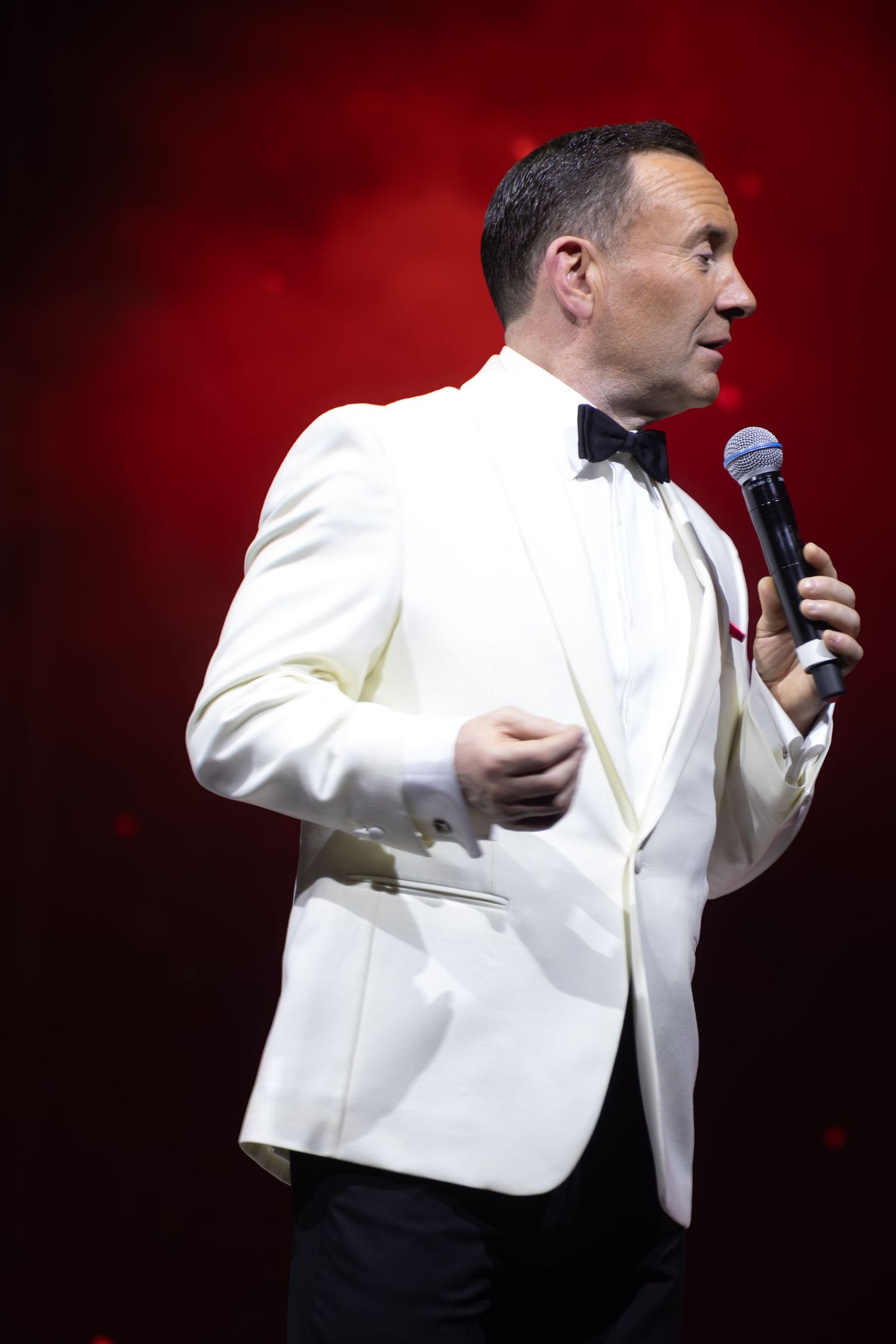 A performer in a white tuxedo jacket sings passionately into a microphone against a red backdrop.