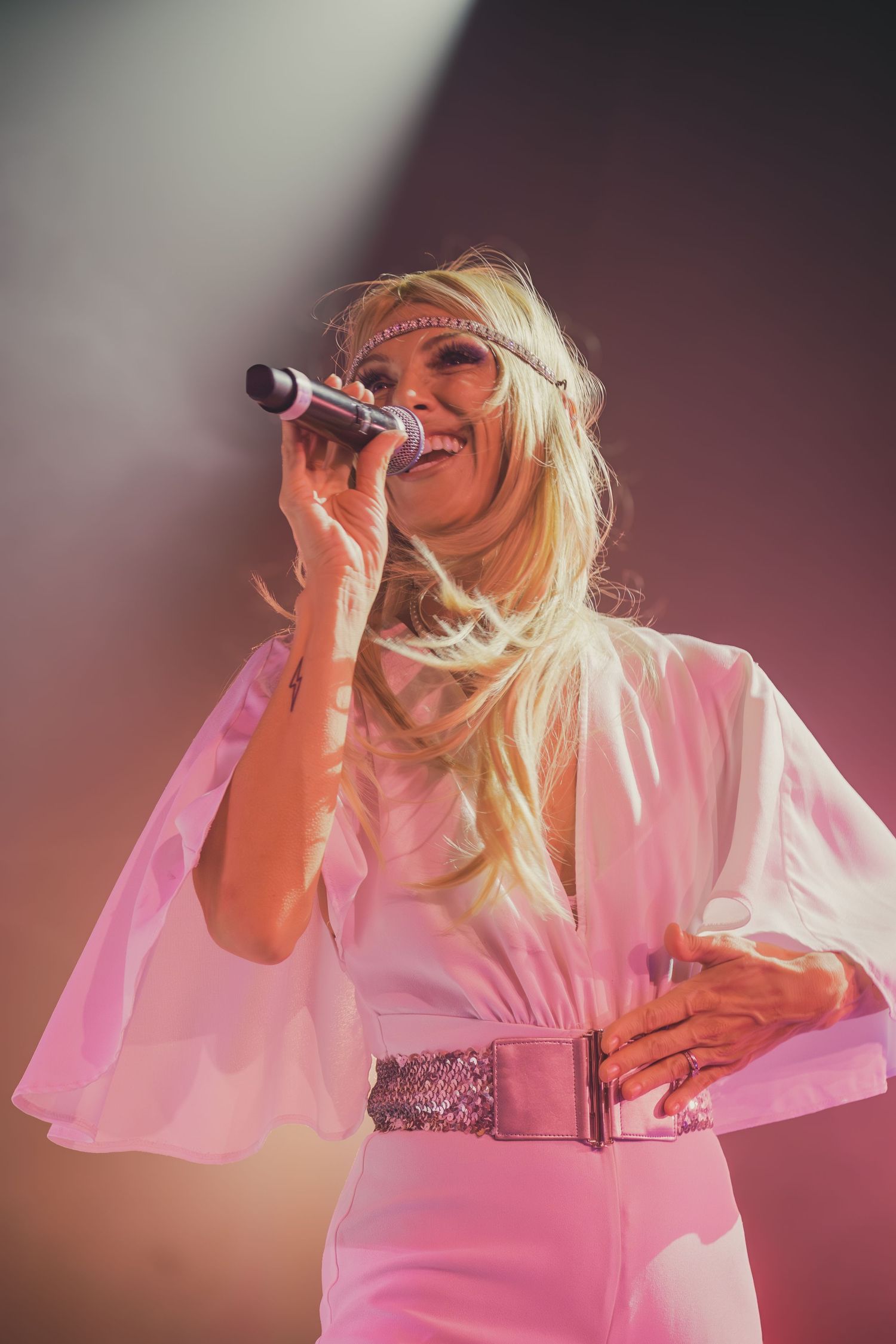 Performer in white dress with silver belt sings passionately under pink concert lighting.