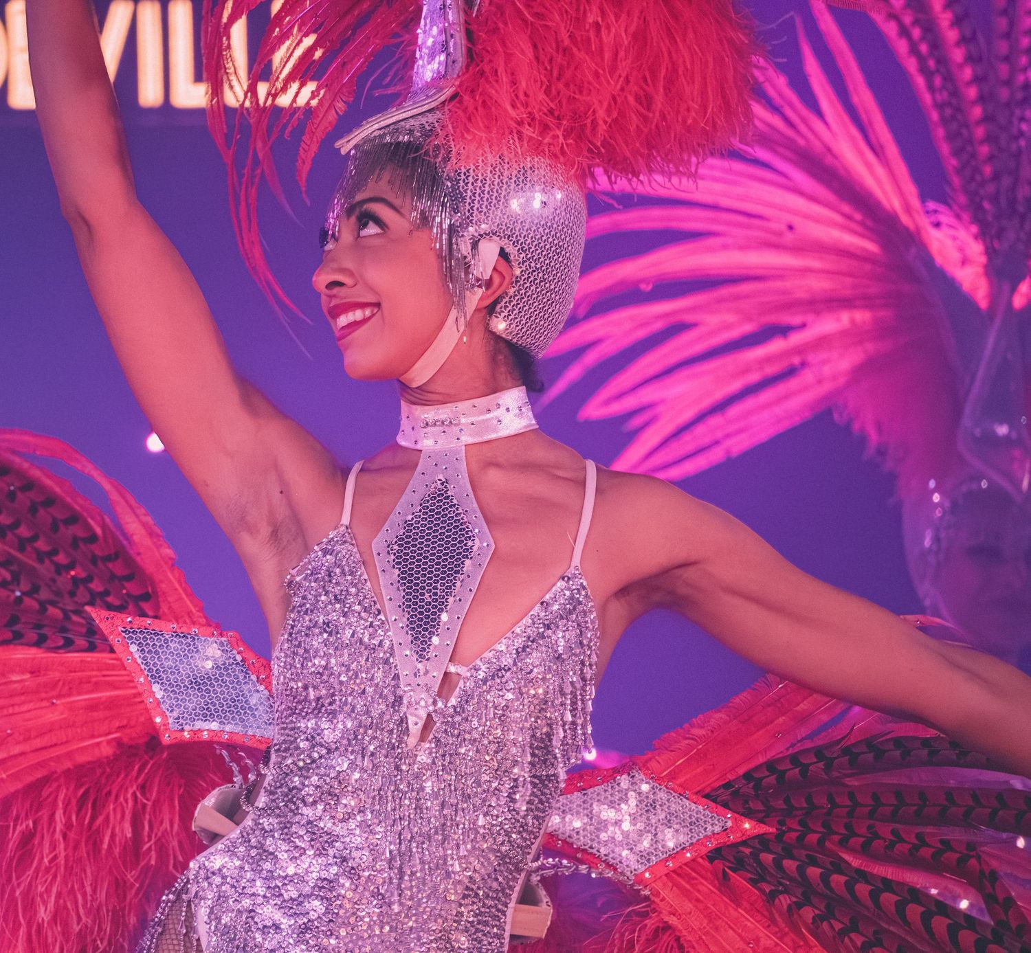 Showgirl in glittering costume with red feather headdress dances under stage lights.