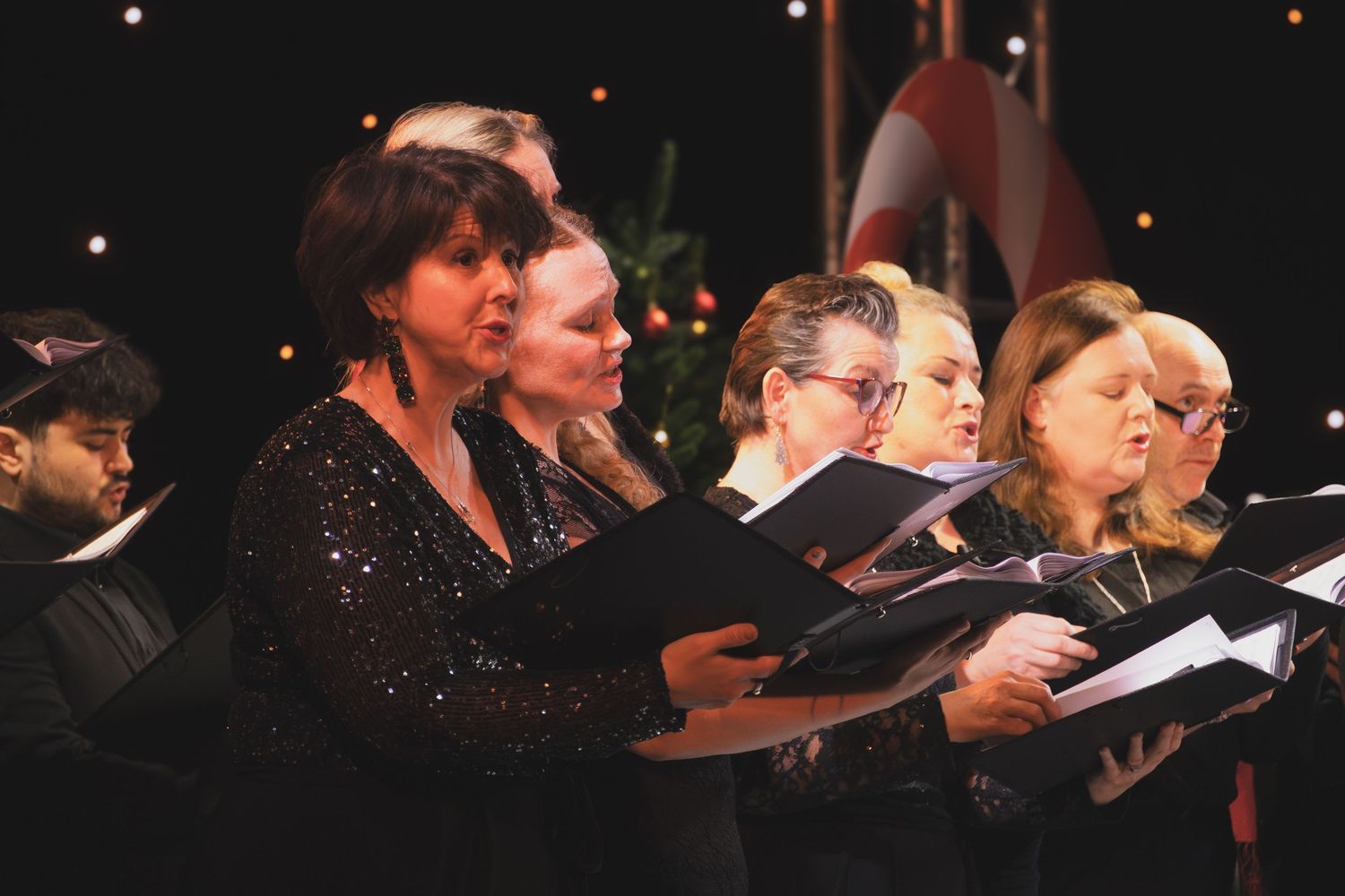Performers holding sheet music stand in a dark concert hall during a choral performance.