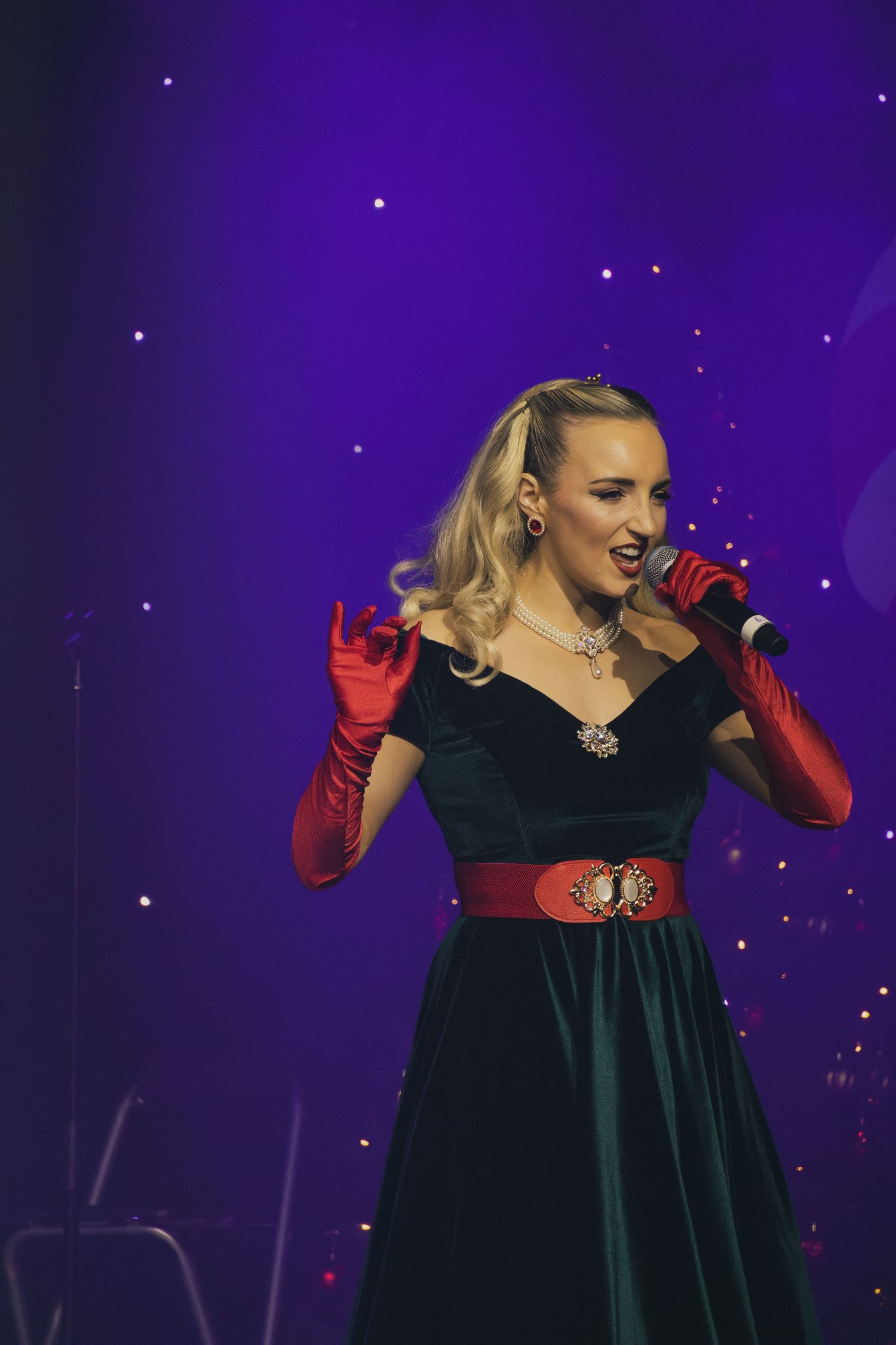 A performer in a green velvet dress with red gloves sings on stage with a purple starlit backdrop.
