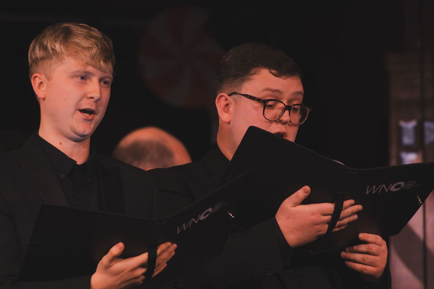 A group of performers in formal attire reading from scripts during a stage performance in dim lighting.
