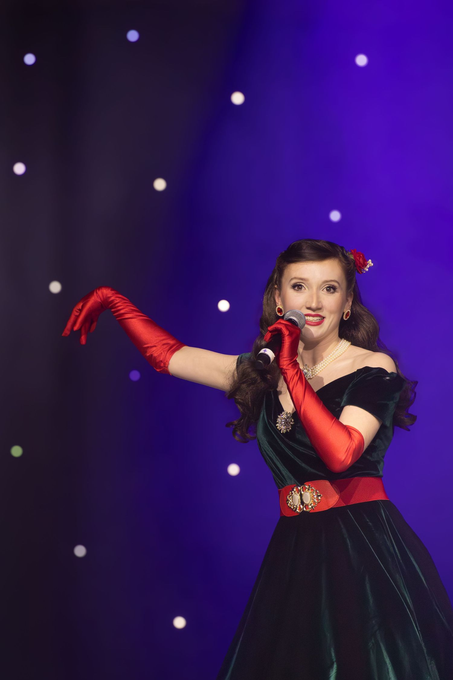 A performer in a vintage-style dress and red gloves poses dramatically against a purple backdrop.