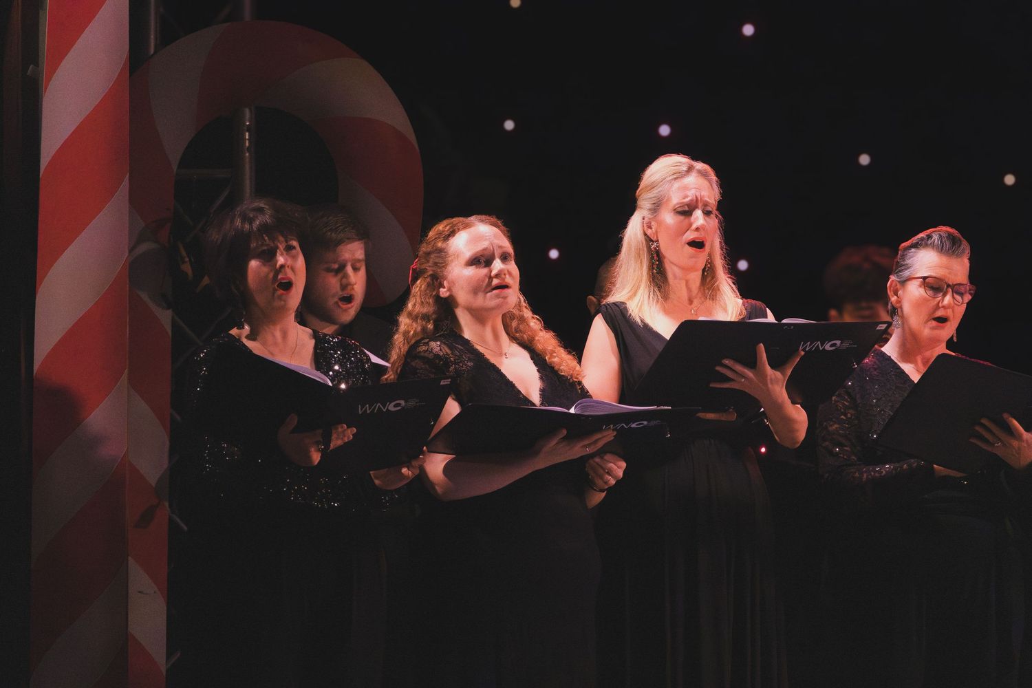 Choir members in formal black attire perform on stage with festive holiday decorations.