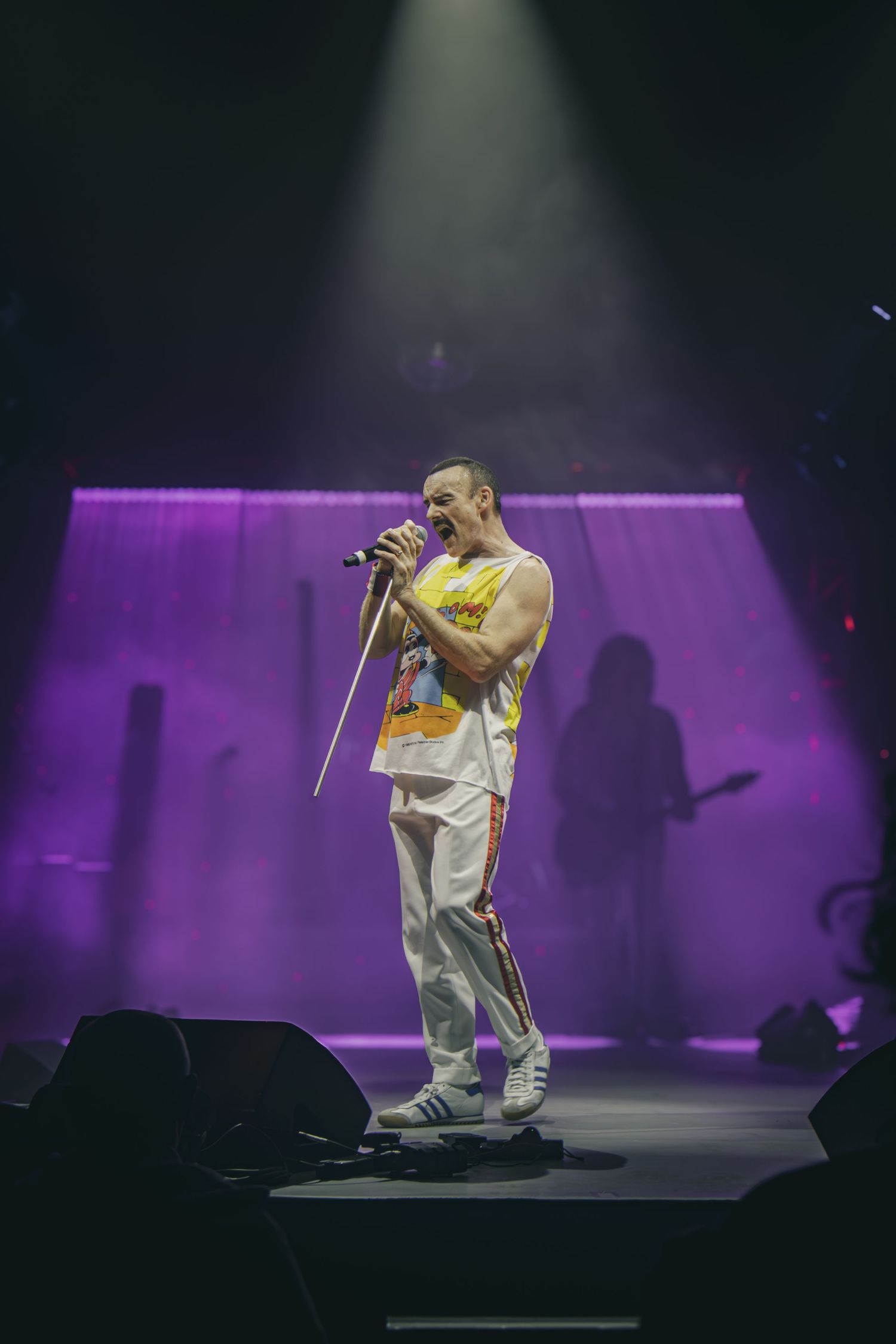 Concert performer in white outfit kneeling on stage with dramatic purple lighting and fog effects in background.