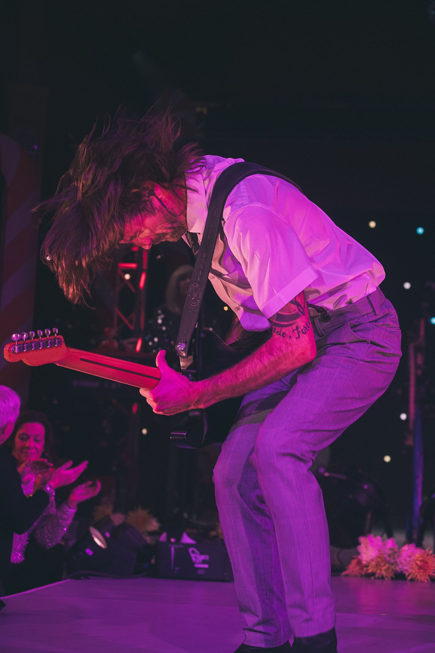 Musicians perform energetically on stage under purple and pink lighting during a rock concert.