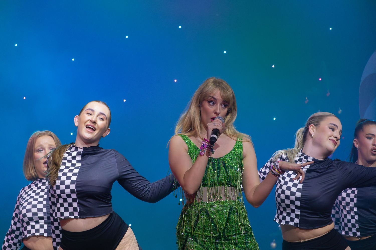 Performance ensemble on stage with mix of green costumes and checkered outfits under blue lighting.