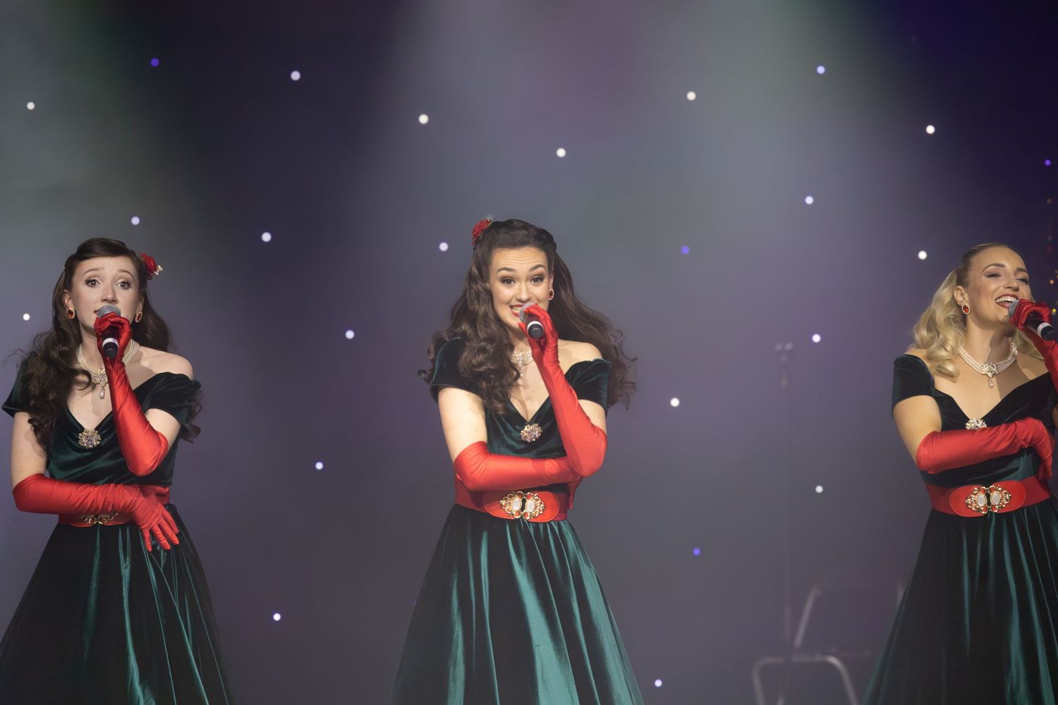 Three performers in matching green dresses and red belts sing on stage with twinkling stars in the background.