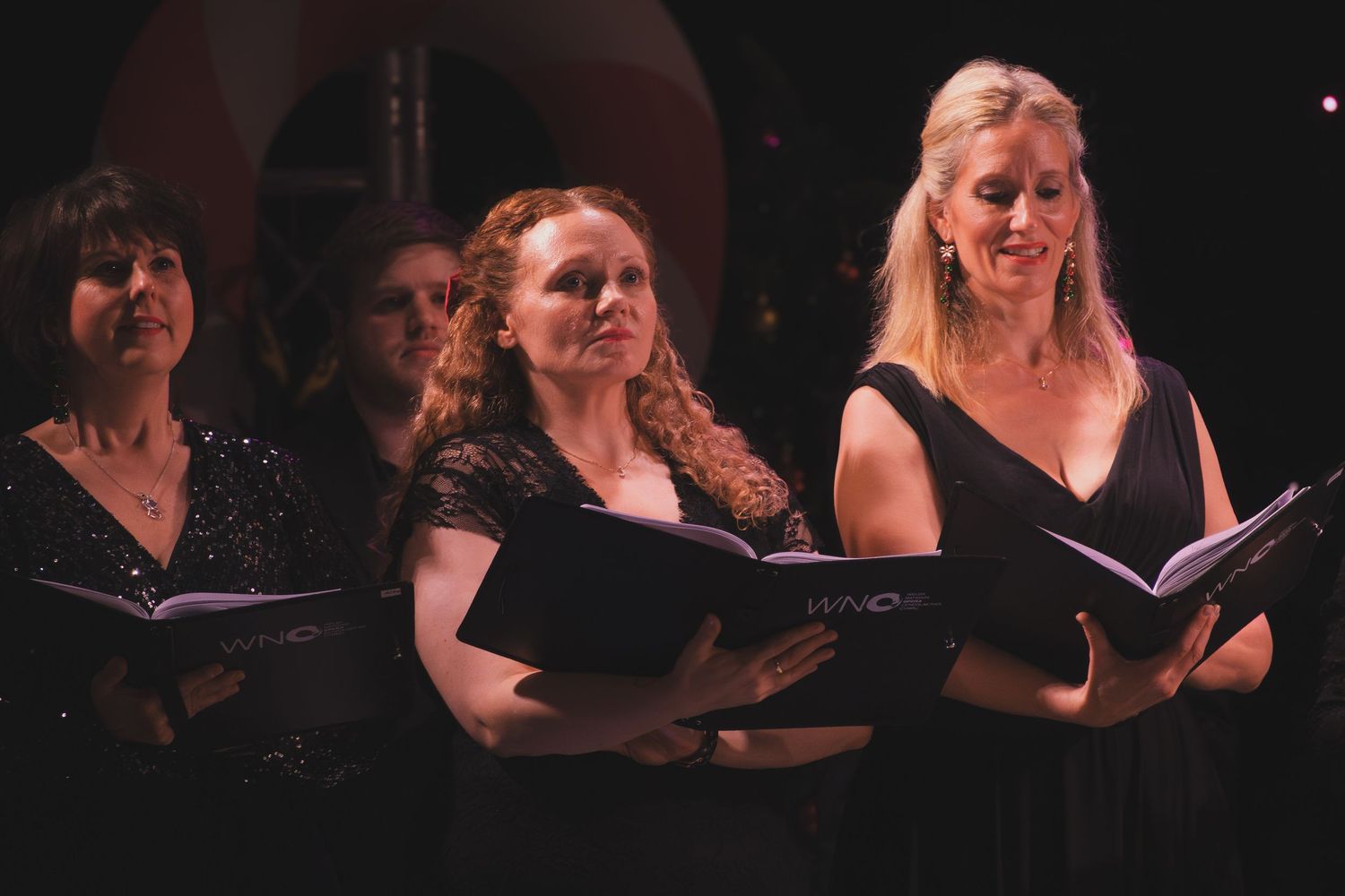 Female choir members in black dresses hold music books while performing on stage.