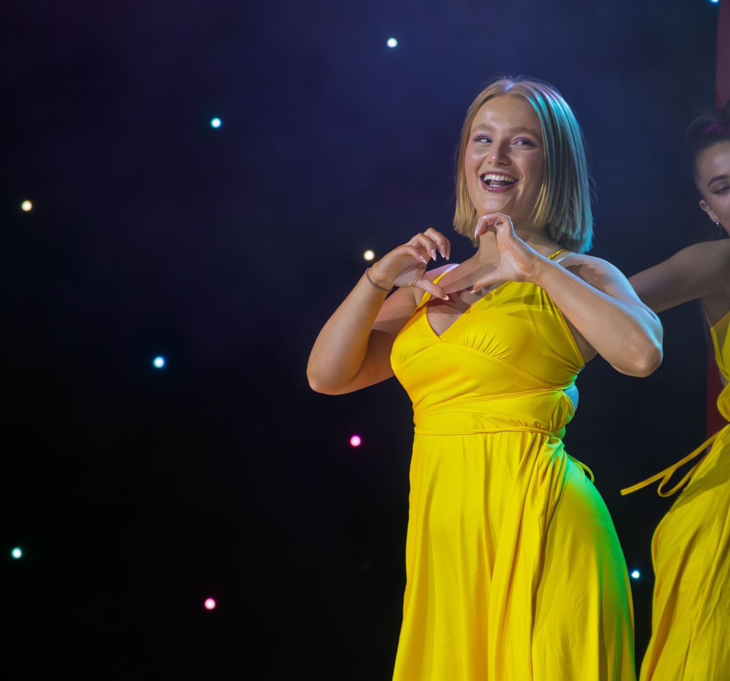 Performer in bright yellow dress singing on stage with twinkling lights in background.