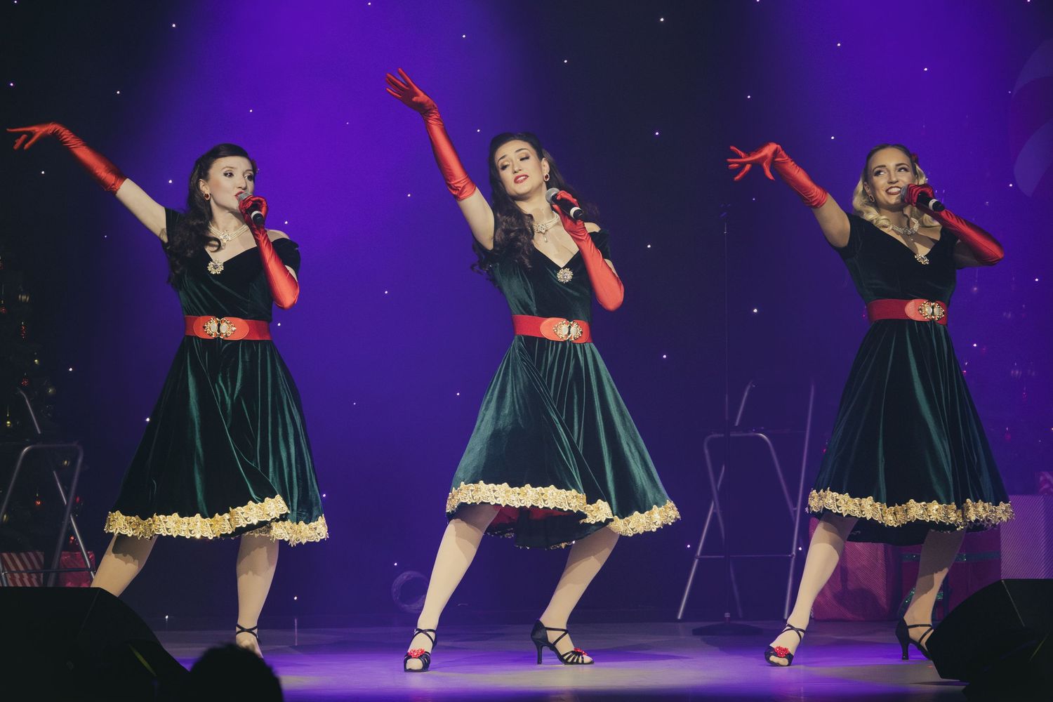 Three dancers in matching green dresses perform synchronized choreography on stage.
