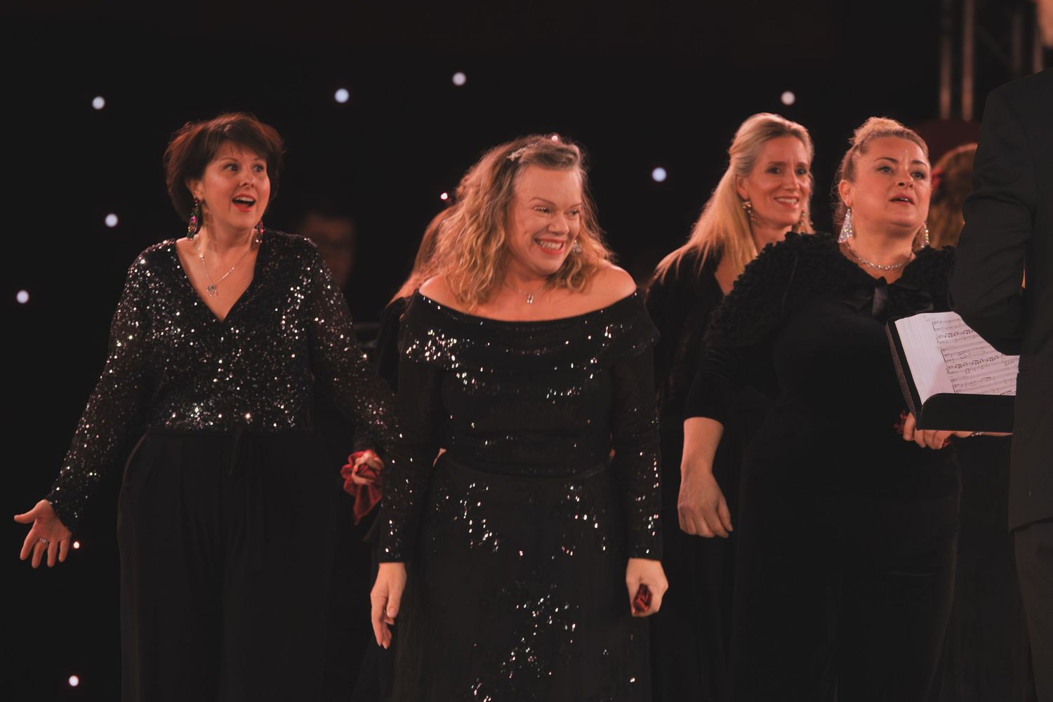 Four performers in black attire sing together on stage with twinkling lights in the background.