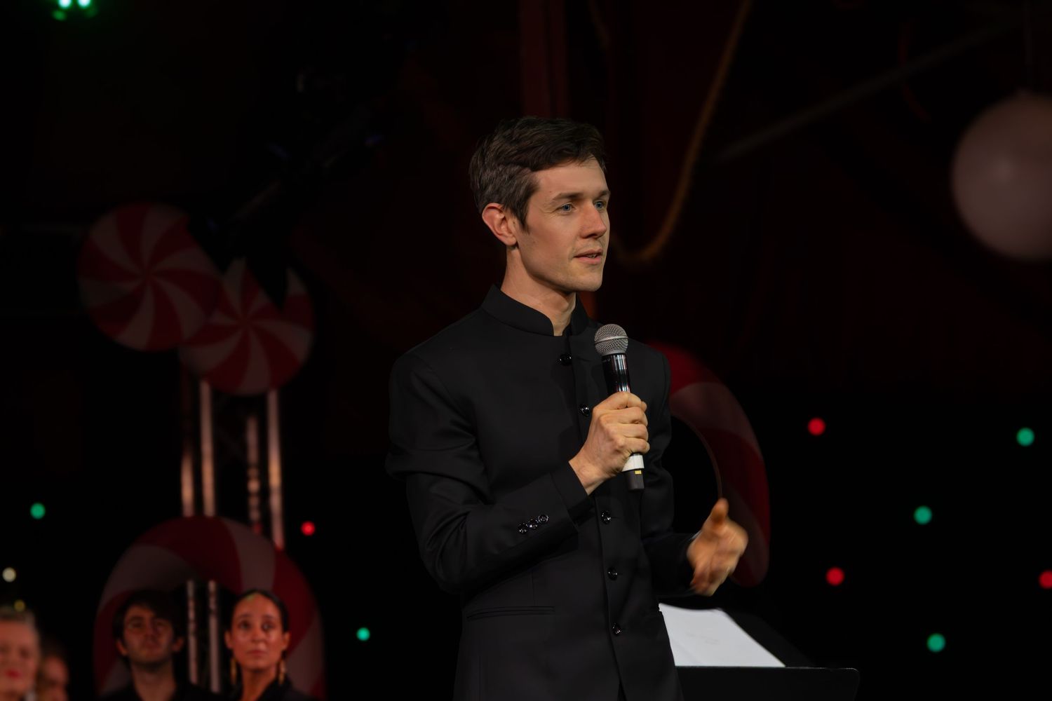 A performer in black attire holds a microphone while singing on stage with colored lights.