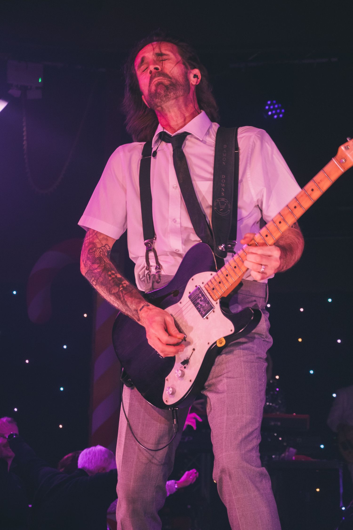 A musician with long hair plays electric guitar while performing in formal attire with suspenders.