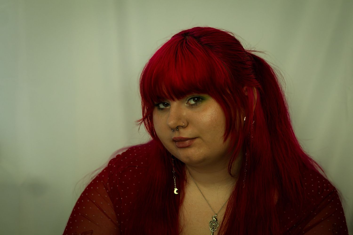 Young woman with long red hair and bangs wearing a red top and necklace, posing against a white background.