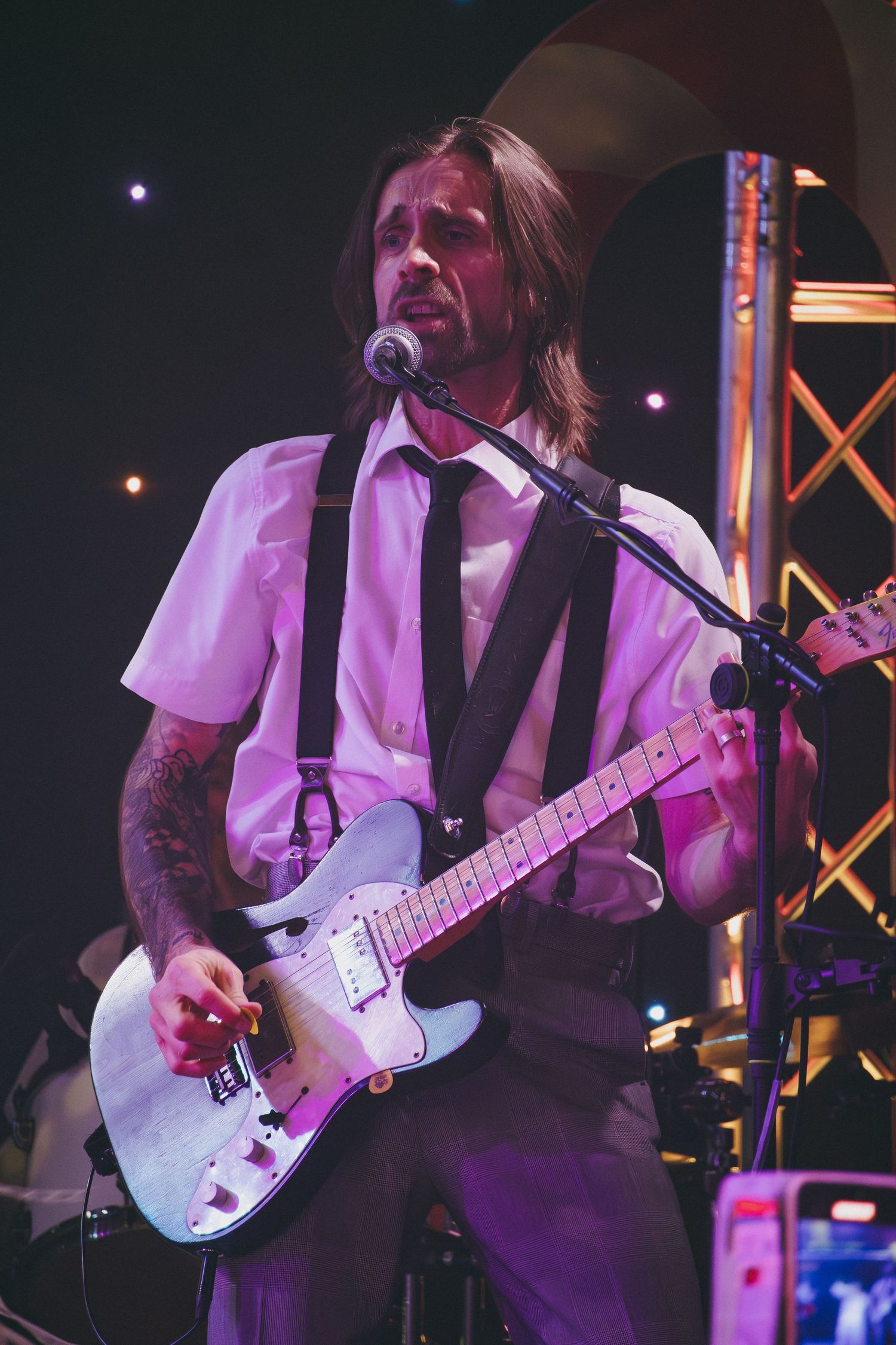 Musician plays electric guitar while wearing white shirt and dark vest under purple stage lights.