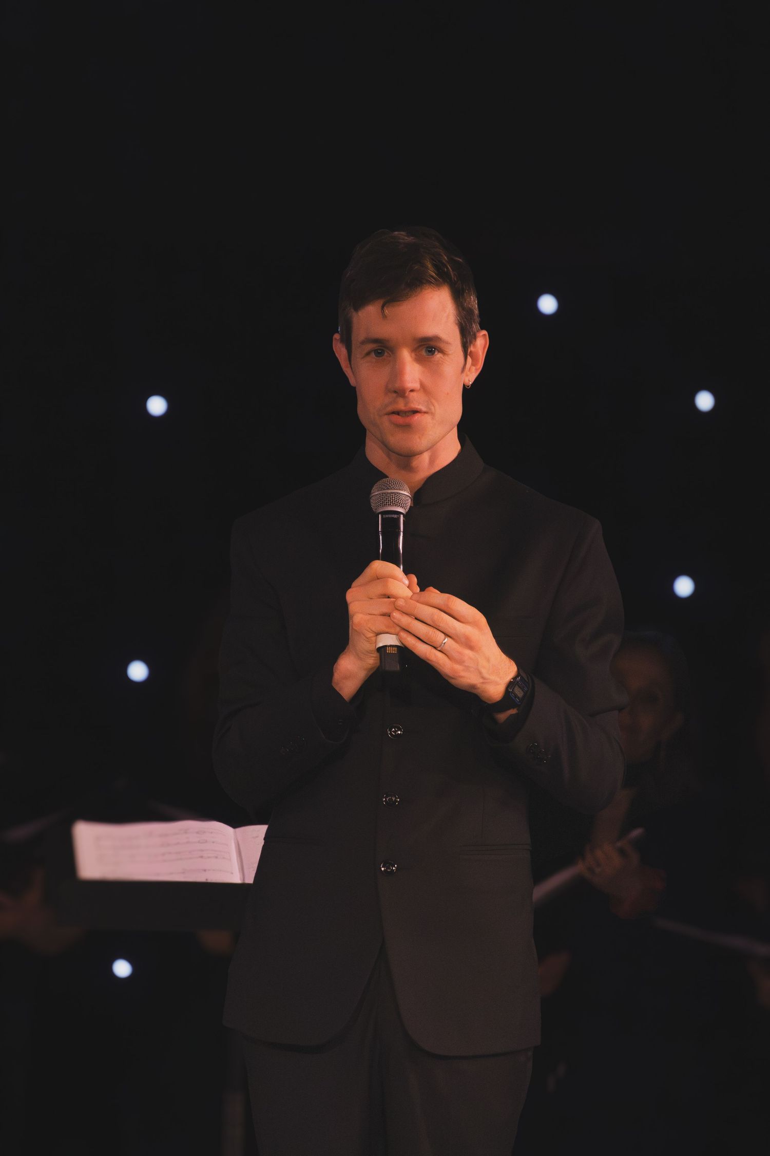 A performer in a black suit holds a microphone on a dark stage with glowing lights behind.