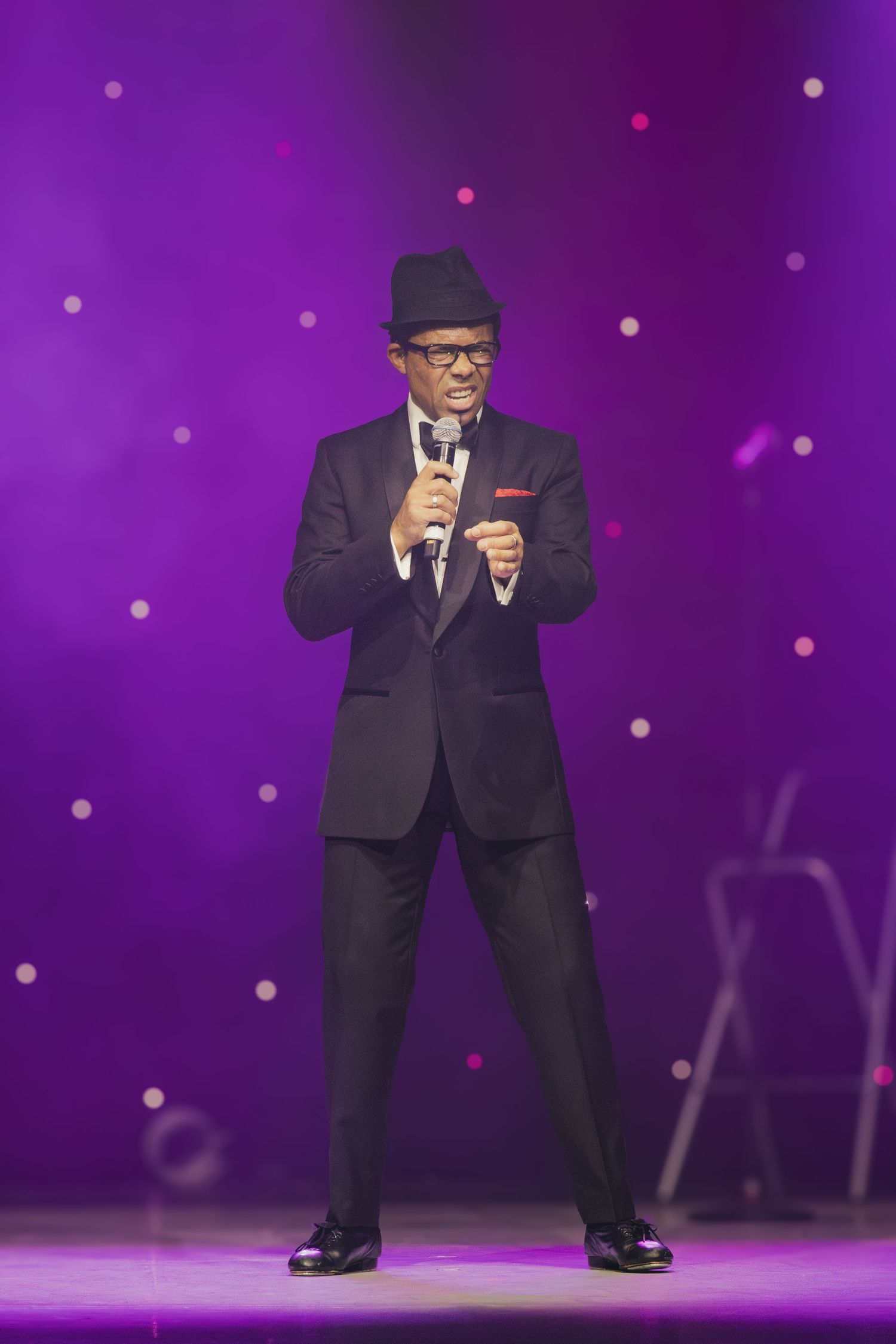Performer in black suit and hat dances energetically on purple-lit stage with sparkly backdrop.