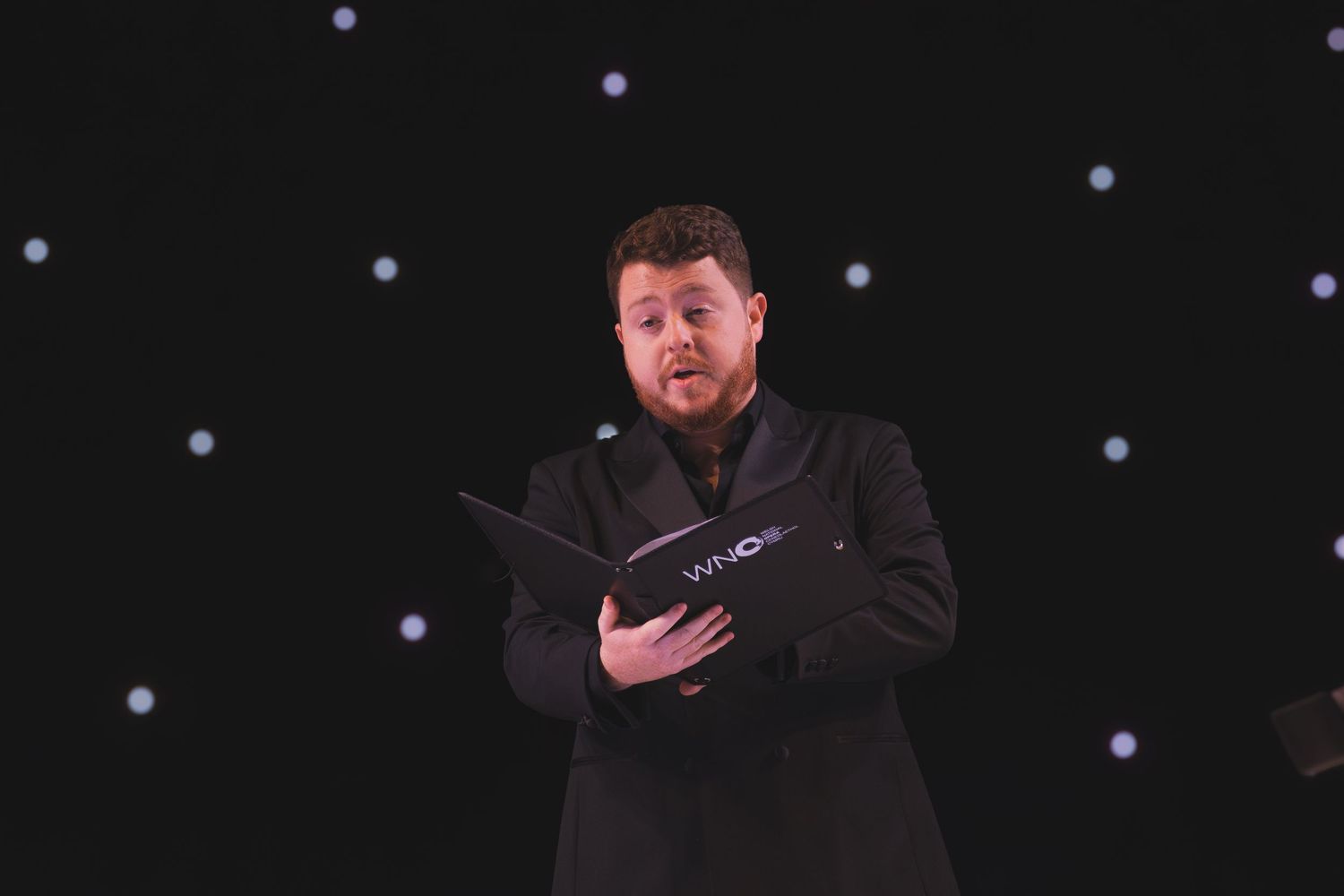 Singers performing in formal black attire on stage with twinkling lights behind them.