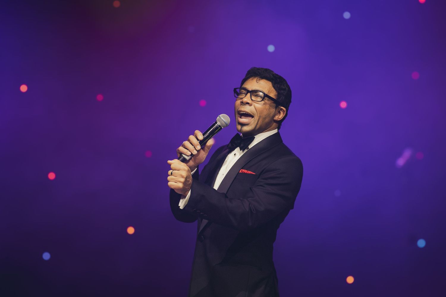 A performer wearing glasses and a tuxedo sings into a microphone against a purple-lit backdrop.