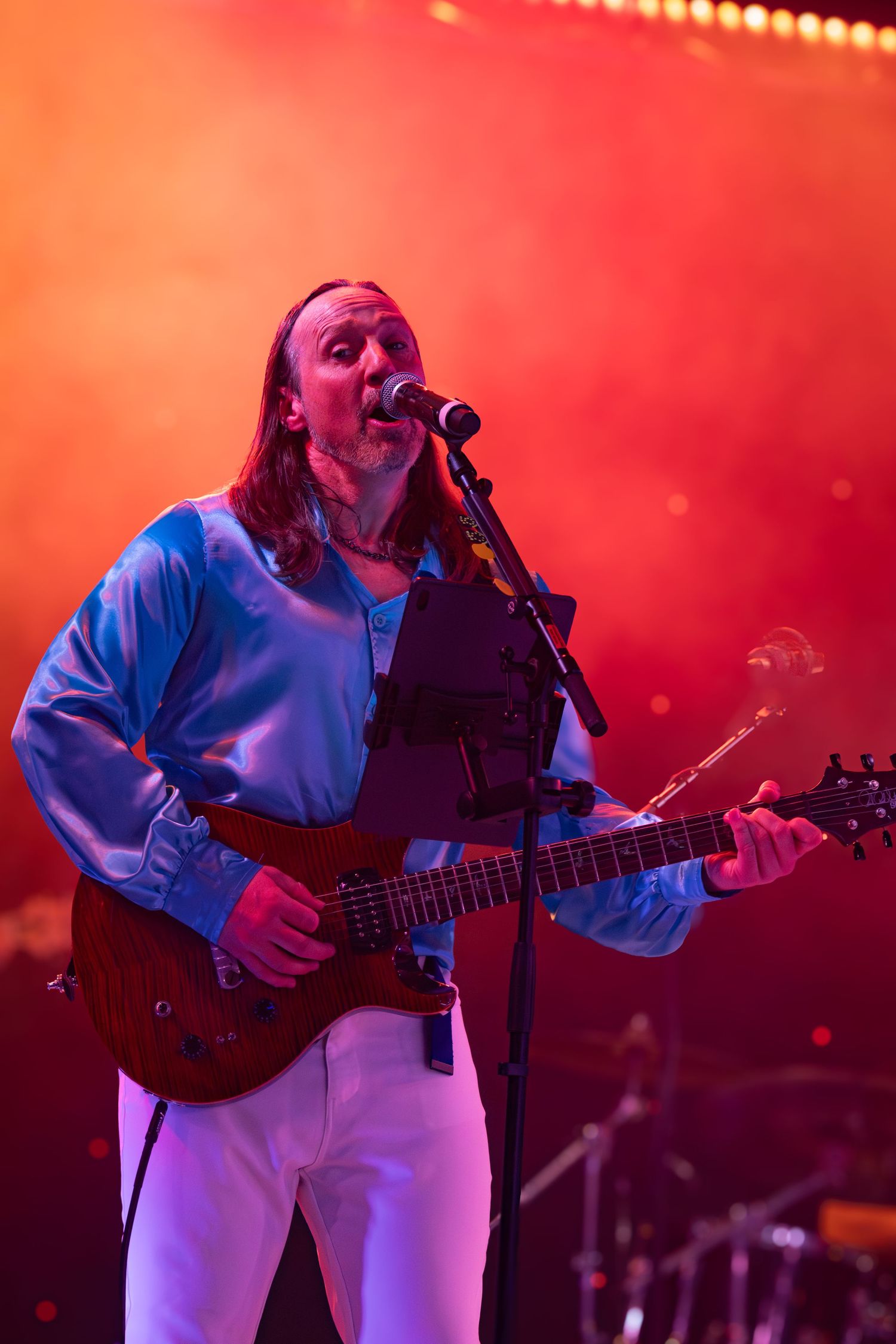 Musician in white outfit playing guitar during performance with red stage lighting.