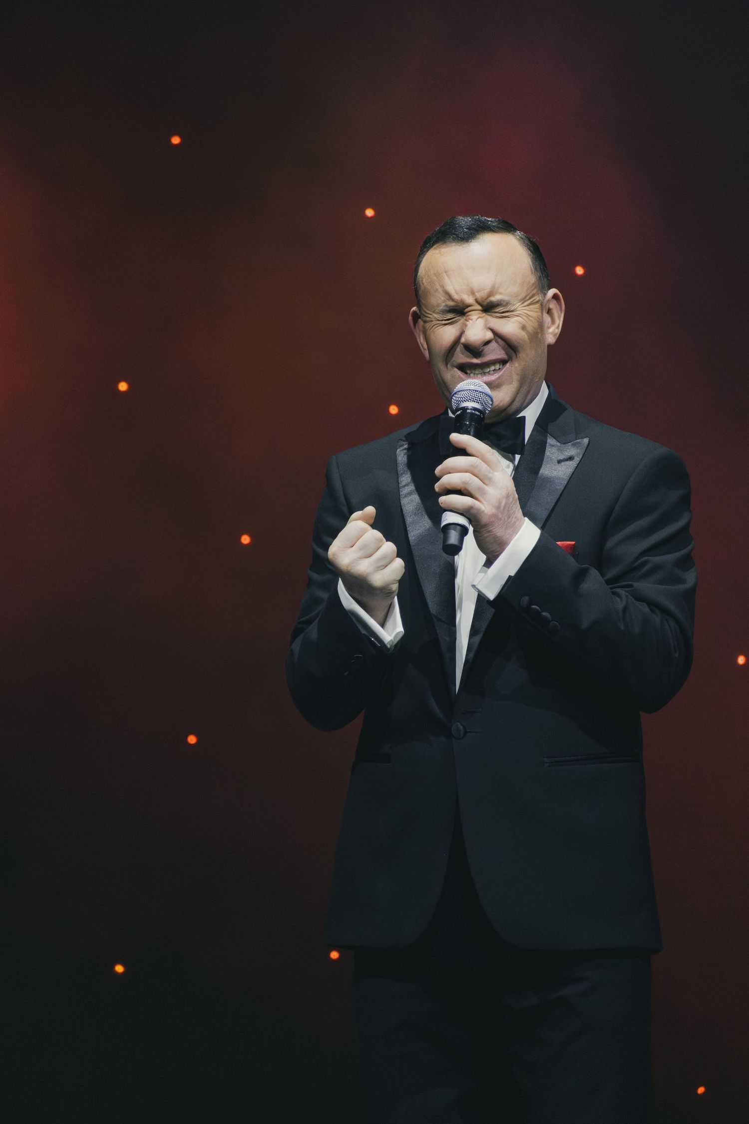 A performer in a black tuxedo sings emotively into a microphone on a dark stage with red lighting.