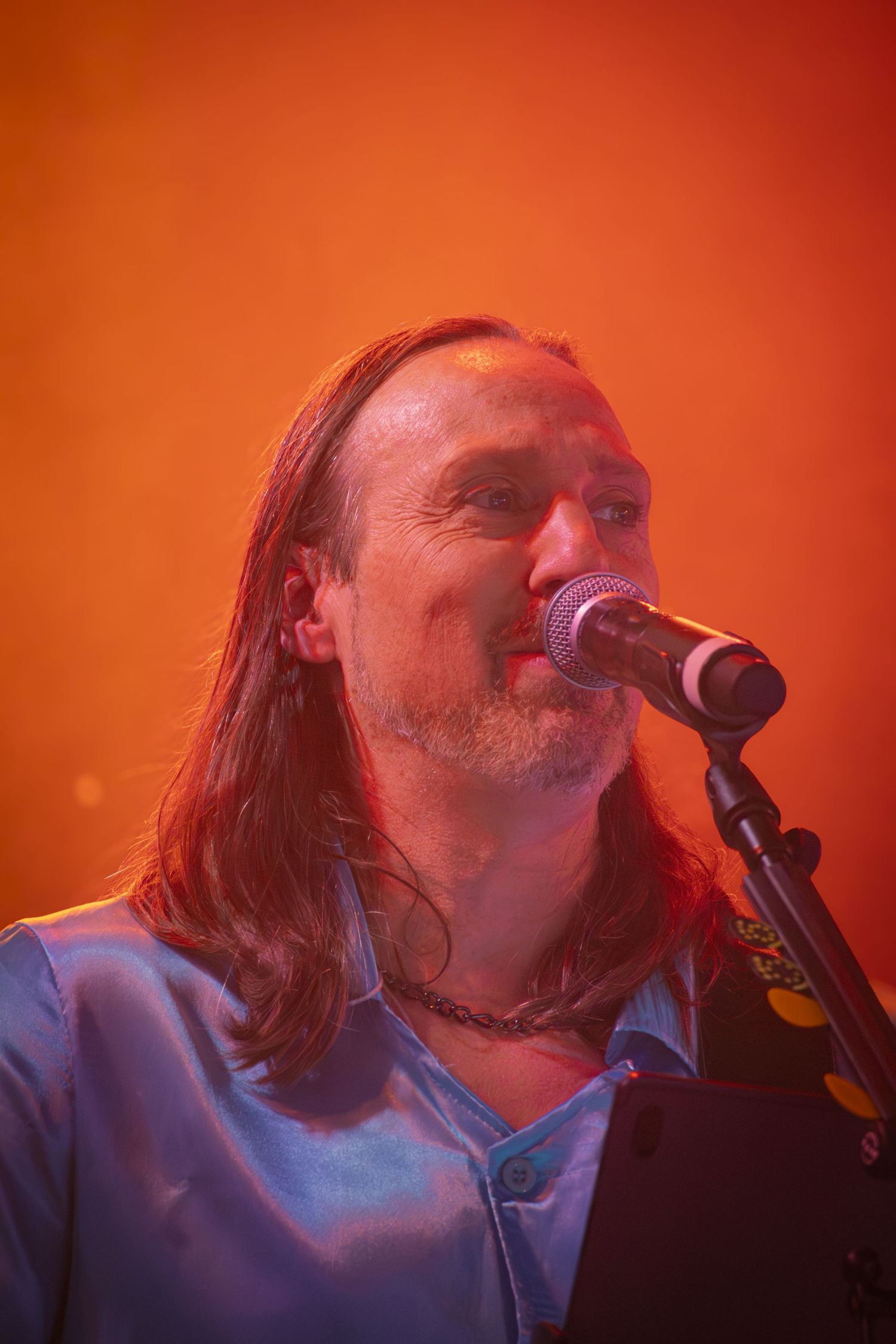 Musician with long hair singing into microphone against orange-red stage lighting background.
