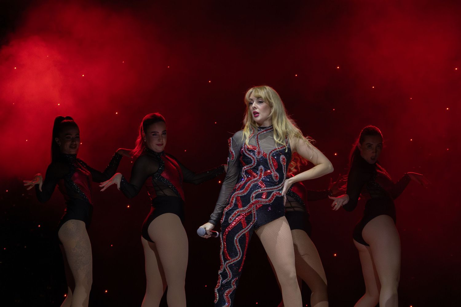 A dance ensemble performs on stage with backup dancers in black outfits against a red backdrop.