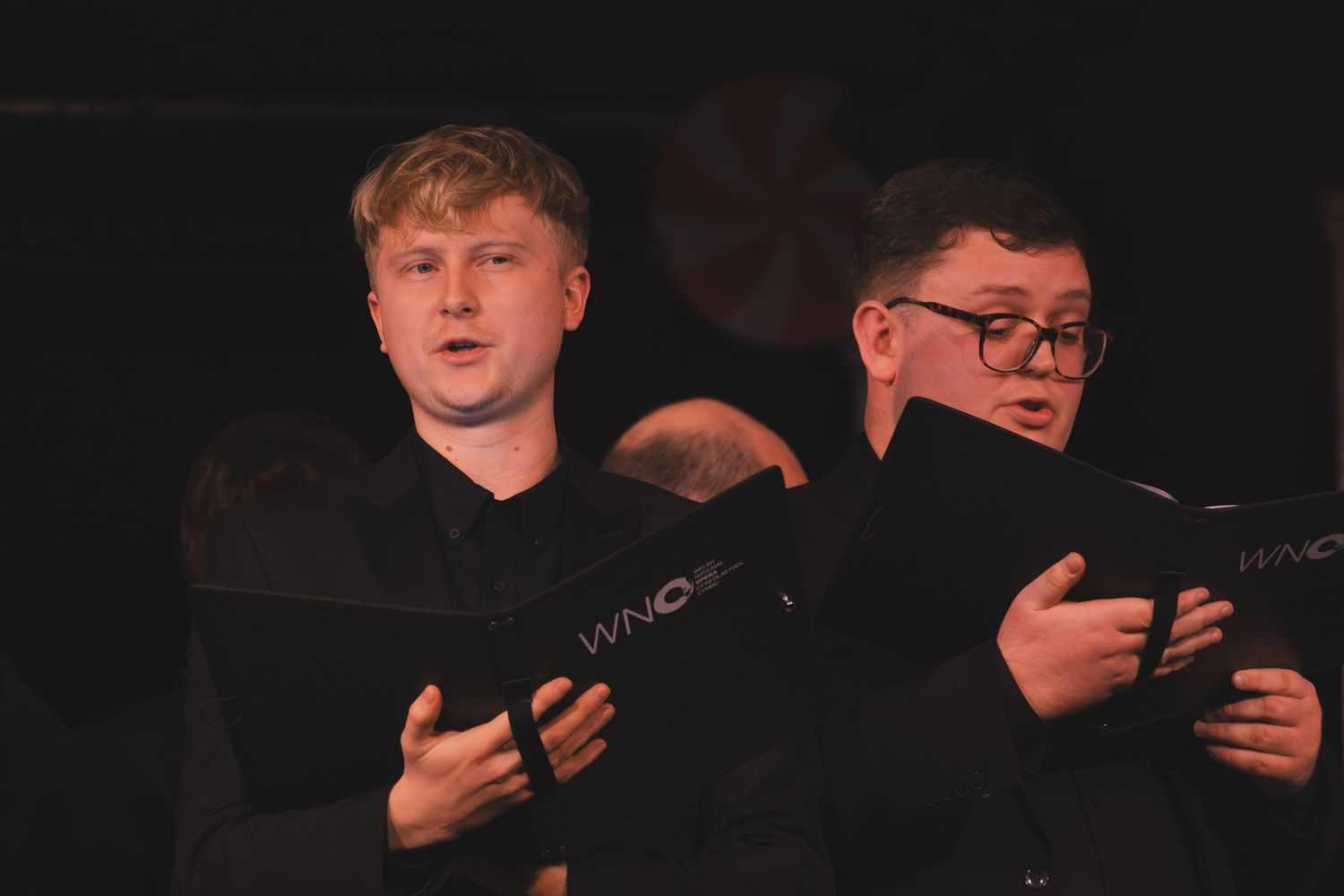 Performers in dark clothing hold music books during a dimly lit choral performance.