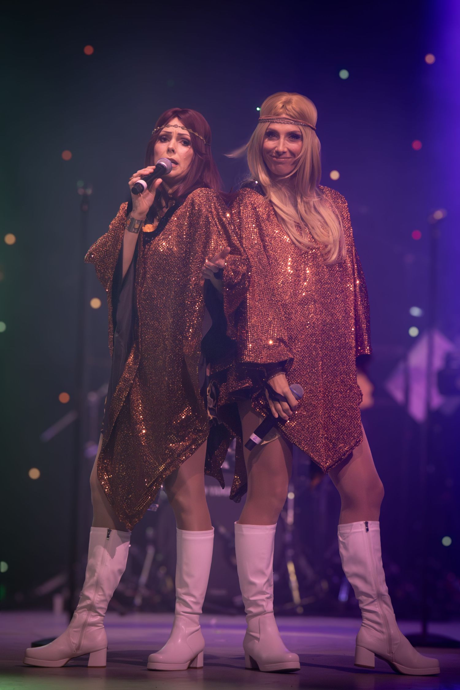 Performers in matching sequined outfits and white boots pose during stage show with colorful lighting.