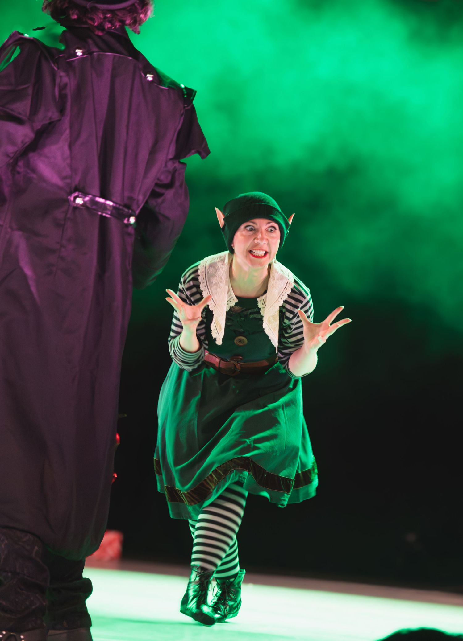 Performer in striped tights and green costume dances energetically against a vibrant green backdrop.