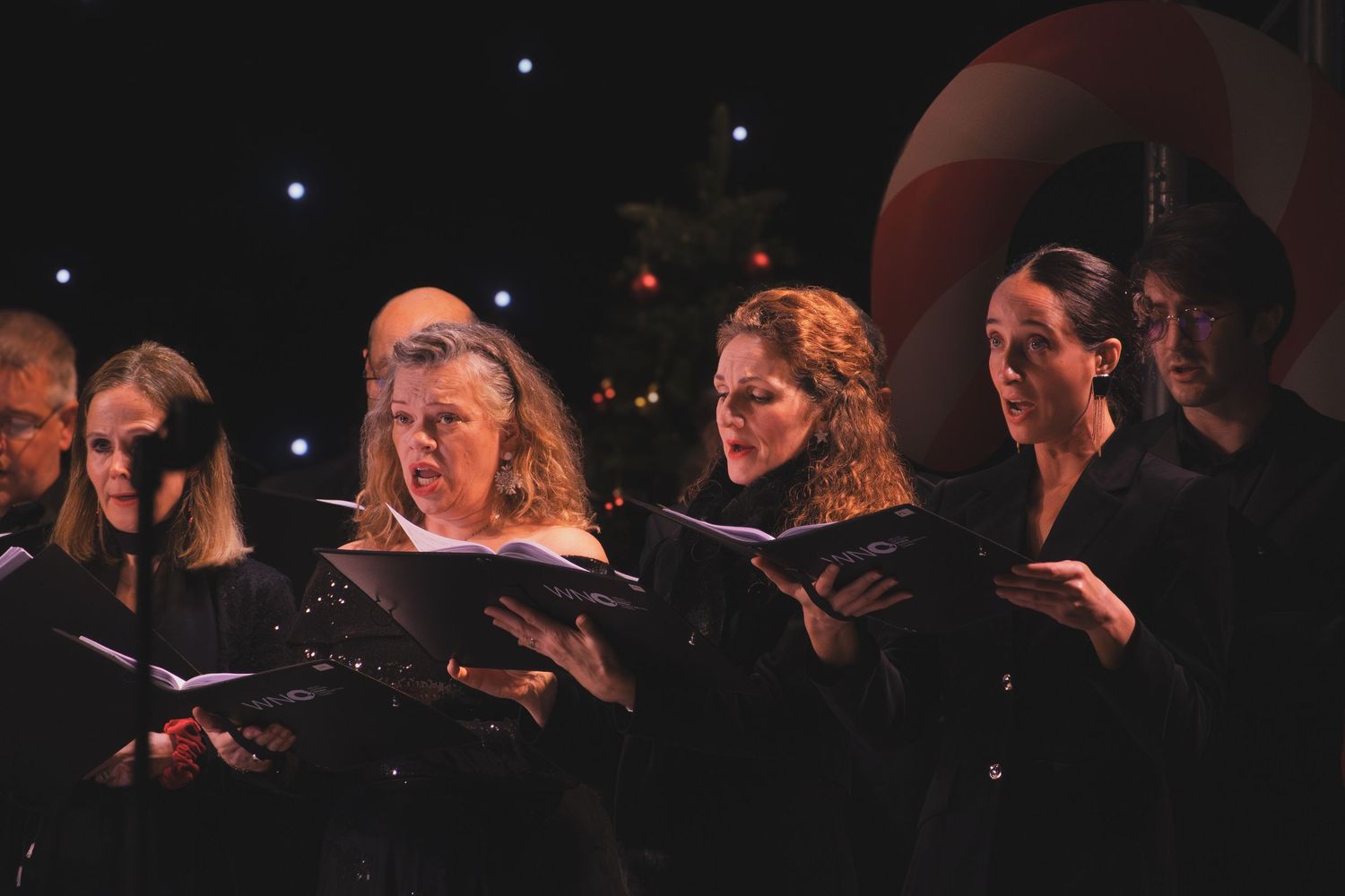 Choir performers in black attire sing together while holding sheet music during a concert performance.