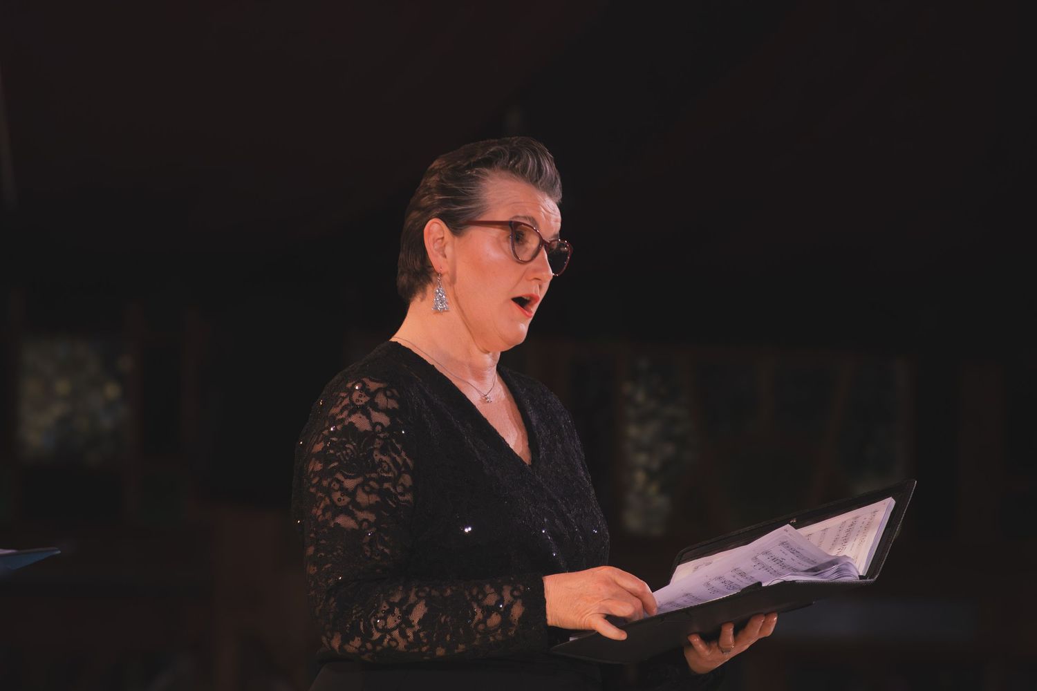 A performer in a black lace dress sings from sheet music during a concert performance.