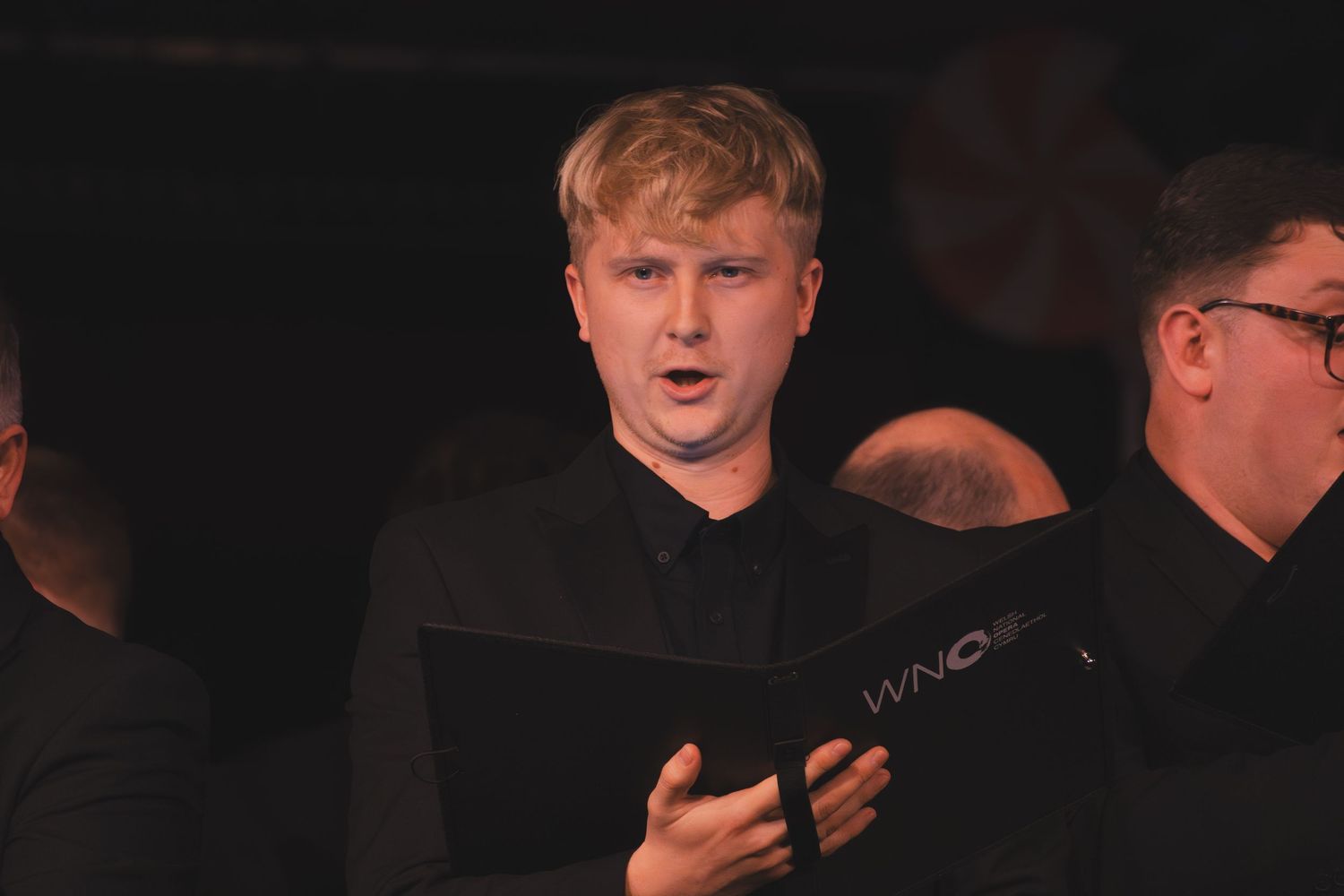 A choir member in black attire sings passionately while holding sheet music during a performance.
