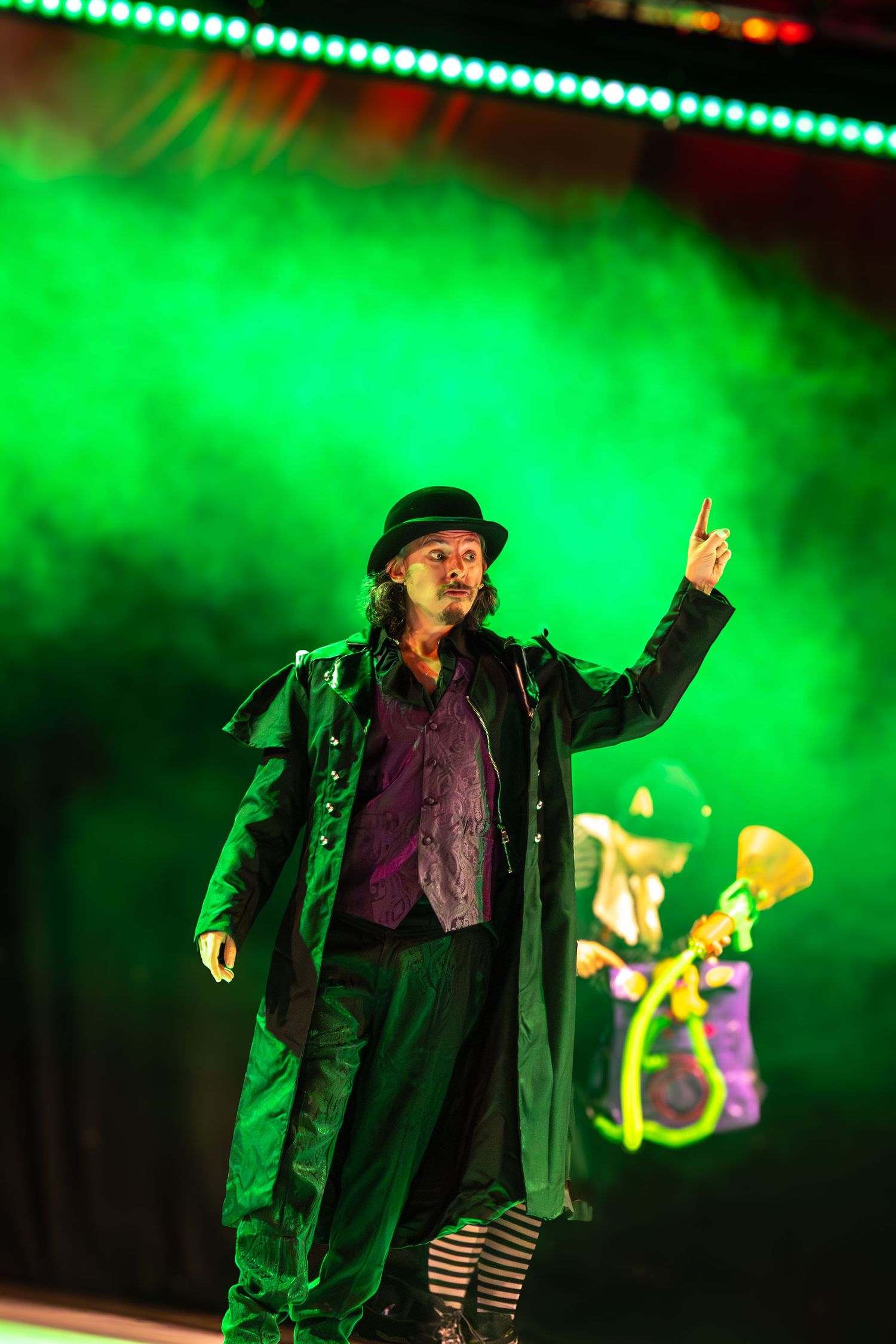 Dramatic performance on stage with figure in black coat illuminated by green atmospheric lighting.