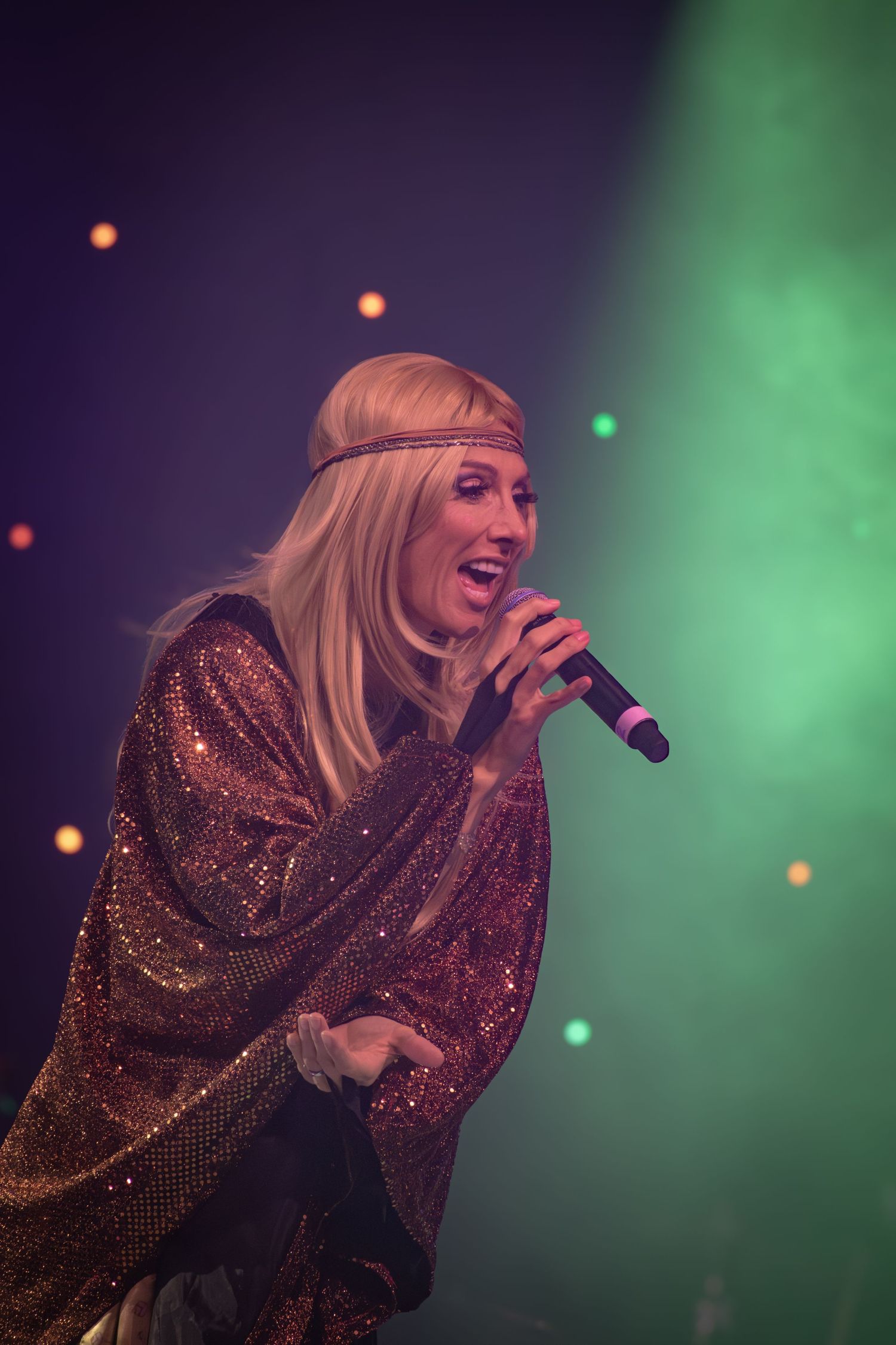 Performer in a glittery dress sings passionately on stage with dramatic purple and green lighting.