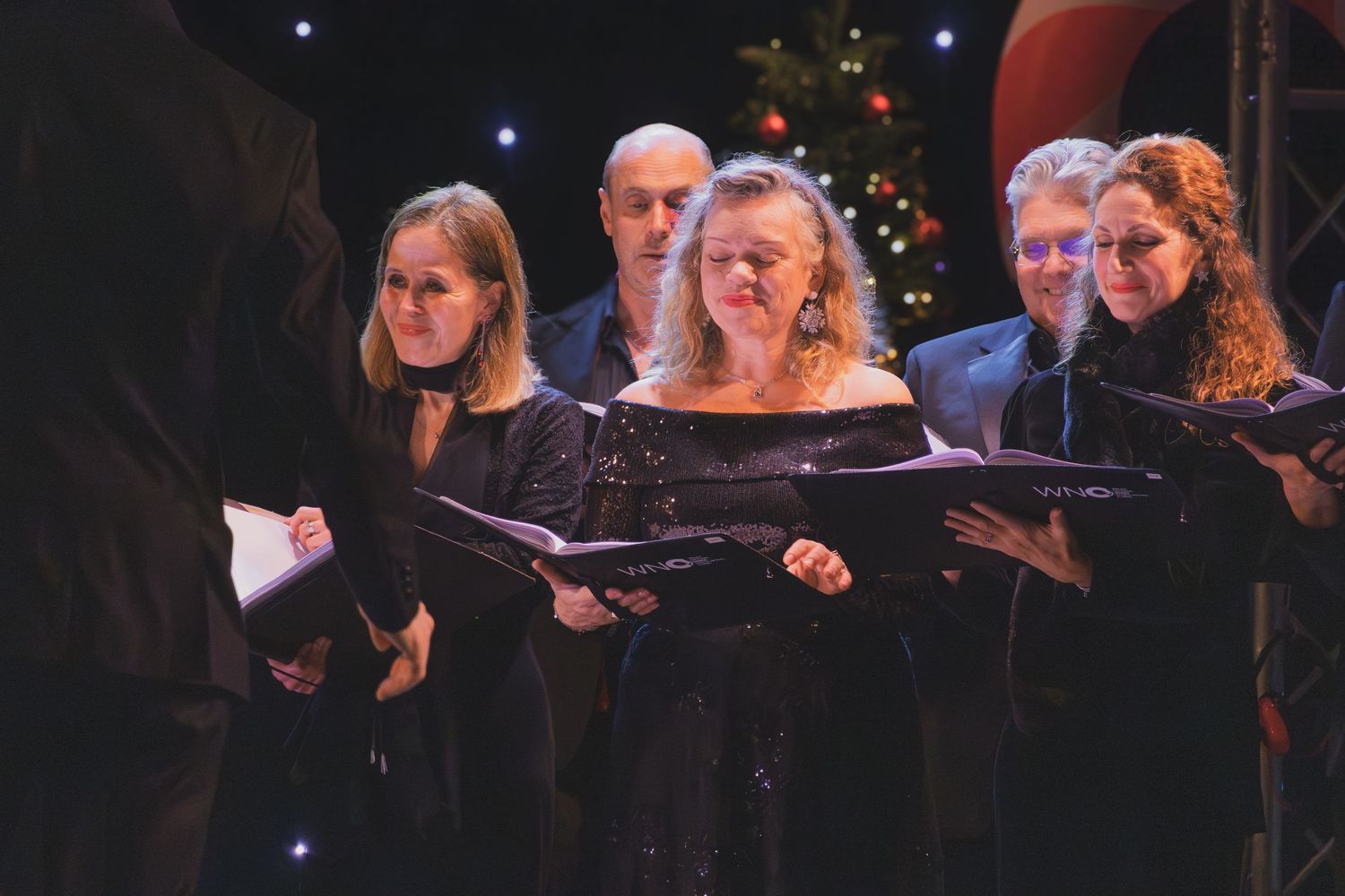 Choir performers in black attire singing together during a holiday concert with twinkling lights in the background.