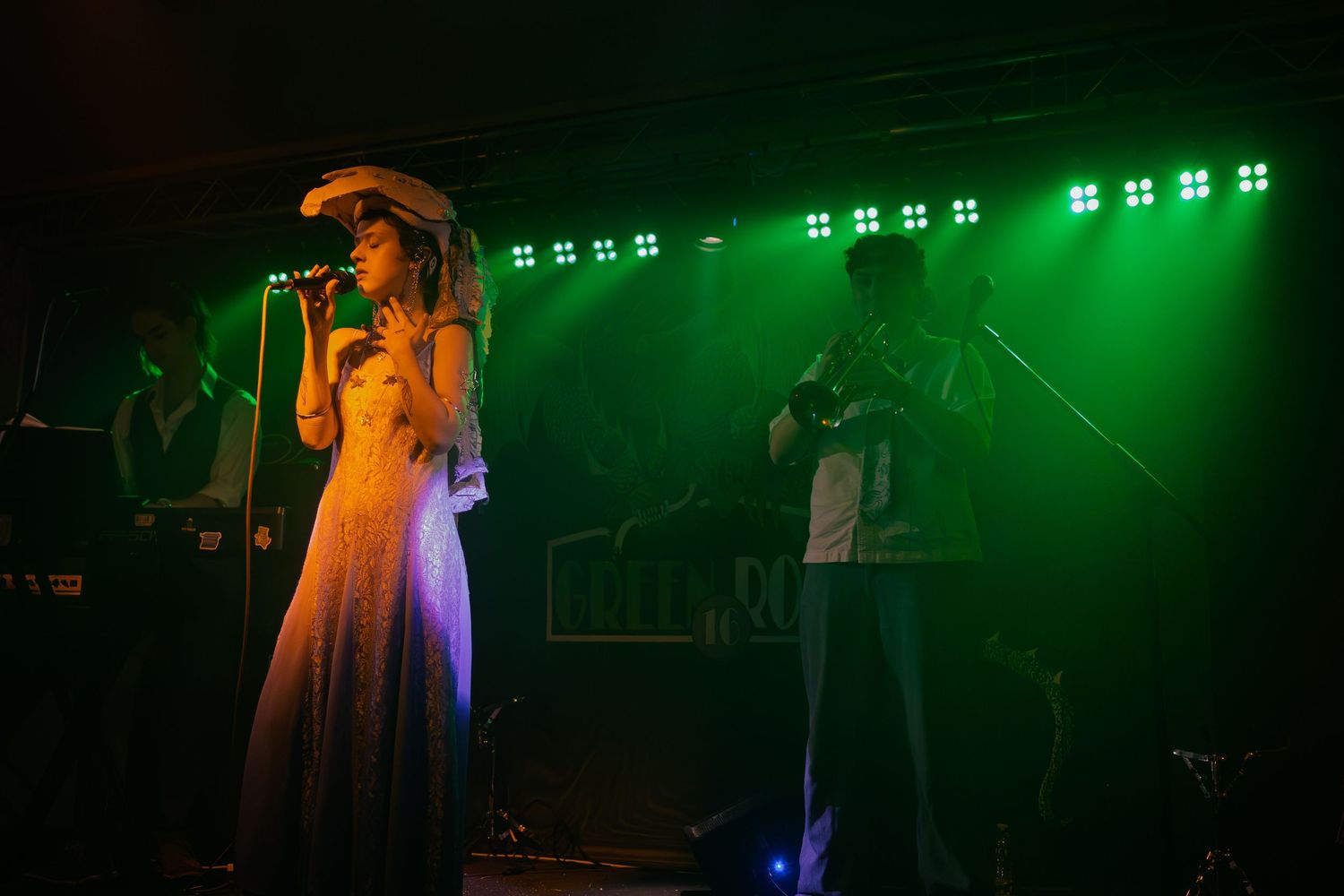 A concert performance featuring a singer in white dress with green stage lighting and band members in background.