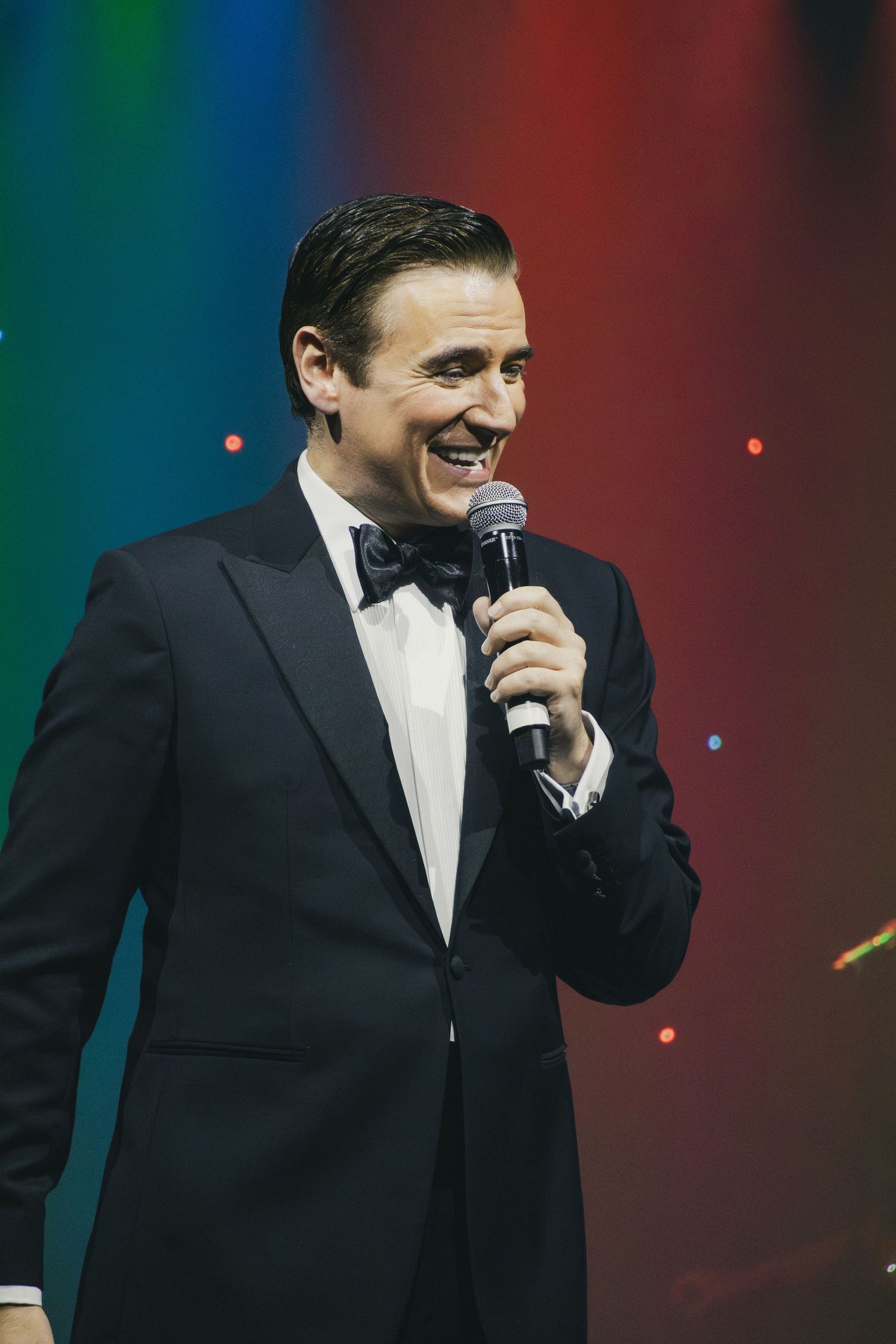 Performer in black tuxedo sings into microphone against colorful stage backdrop.