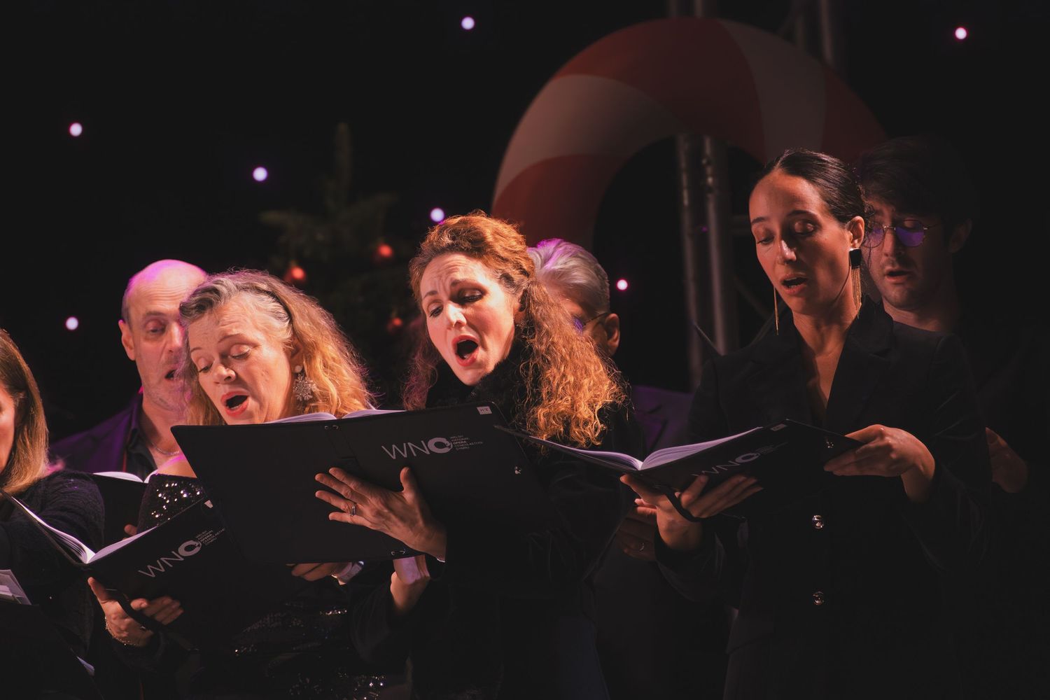 Choir members sing passionately while reading from black music folders during an evening performance.