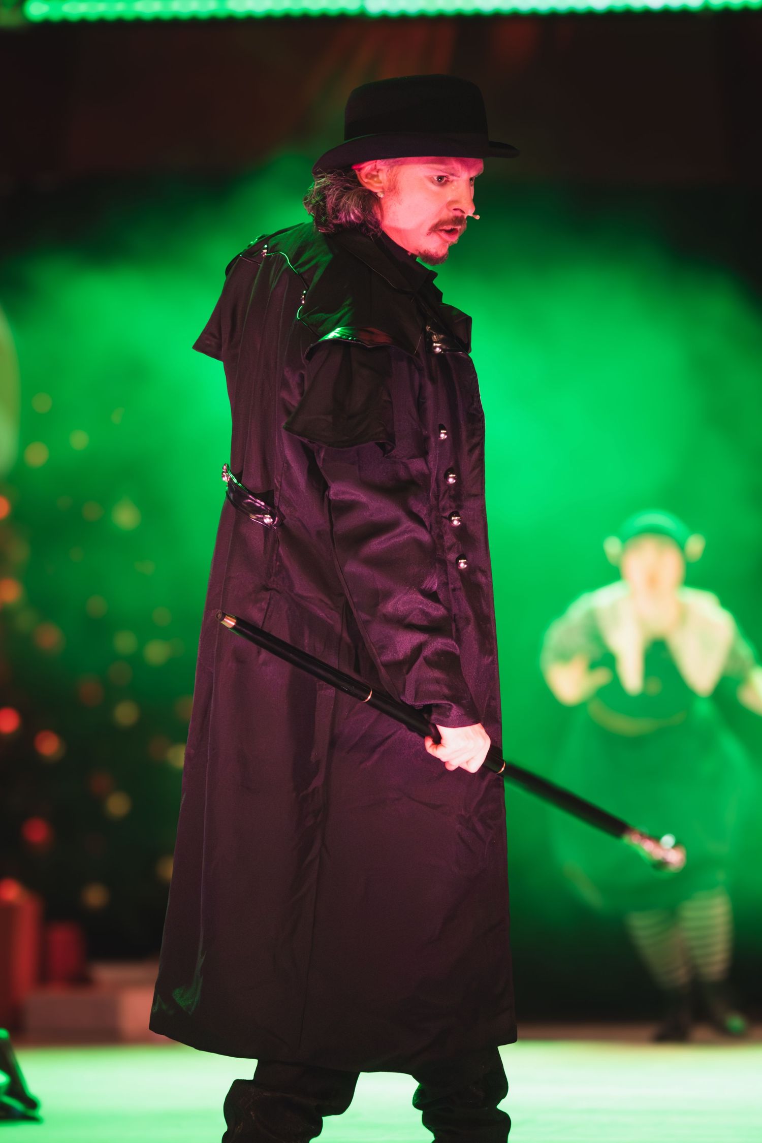 Silhouetted figure in dark coat performs on stage bathed in emerald green lighting.