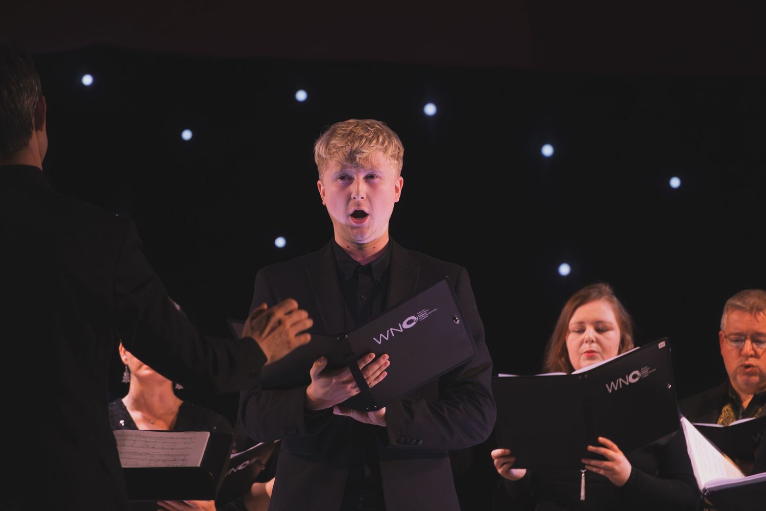 Performers in formal attire sing together on a dark stage with spotlights illuminating them from above.