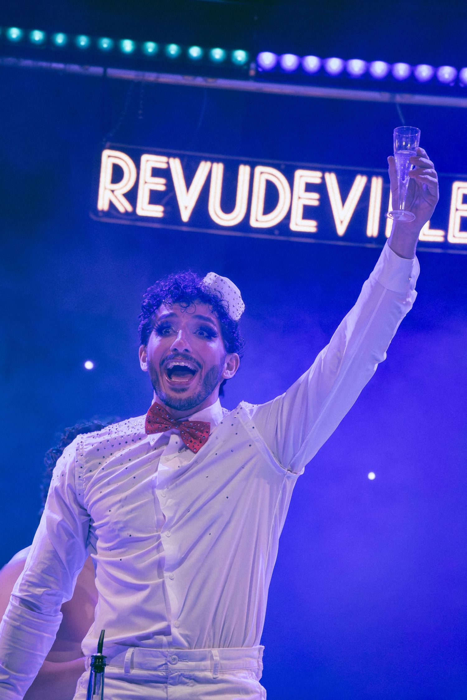 Performers in white shirts dance energetically under blue stage lights with 'Revudeville' text visible.