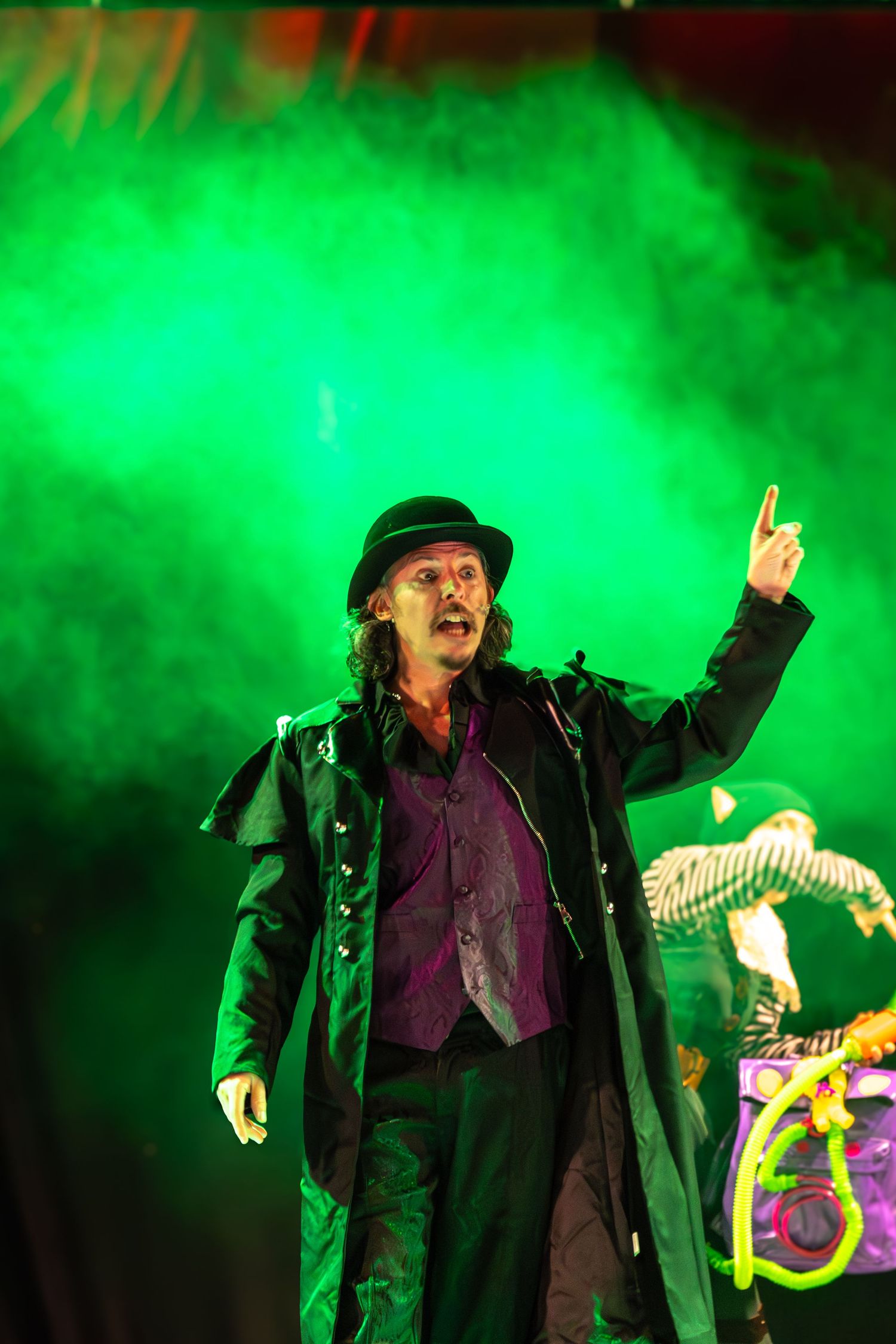 A theatrical performer in black coat and hat gestures dramatically against green fog effects.