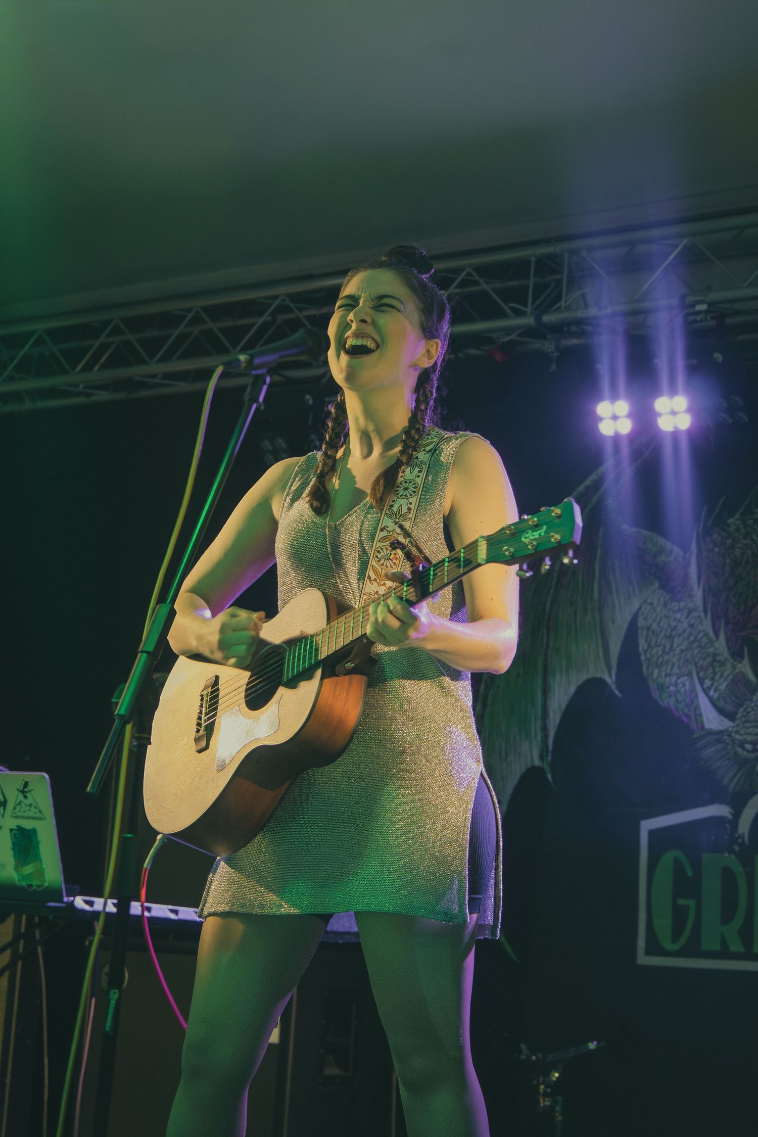 A performer sings passionately while playing acoustic guitar on stage with colorful lighting and sound equipment visible.