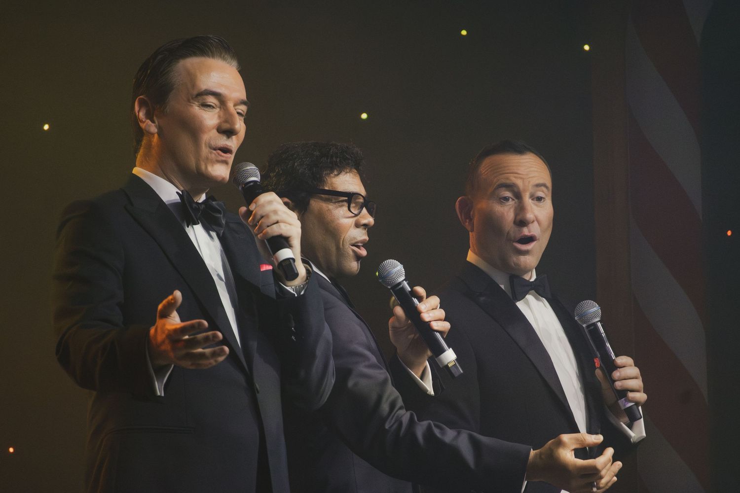 Performers in black tuxedos sing into microphones on a dimly lit stage.